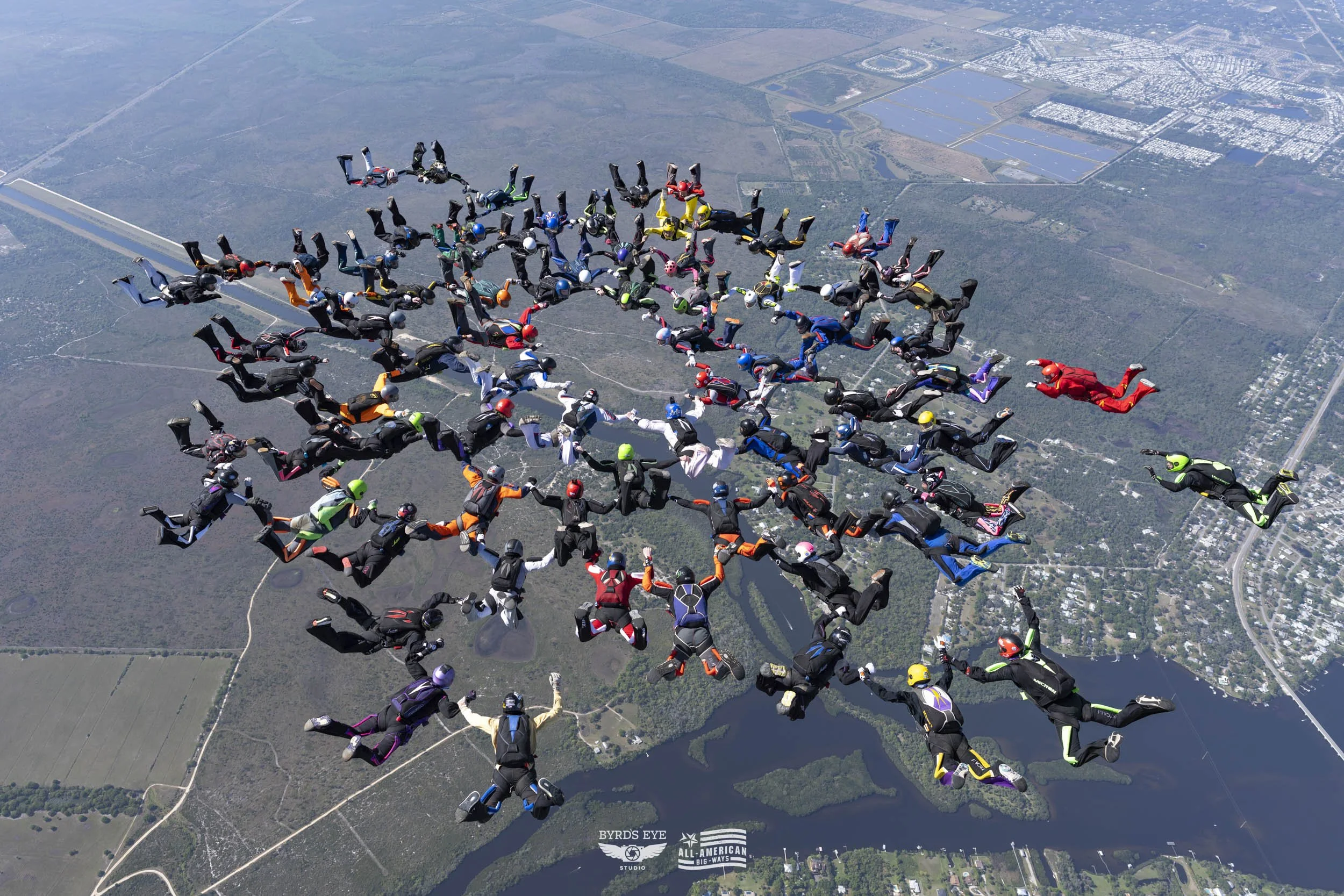 Skydivers in colorful jumpsuits and helmets forming a large group in free fall over a landscape with water and roads.