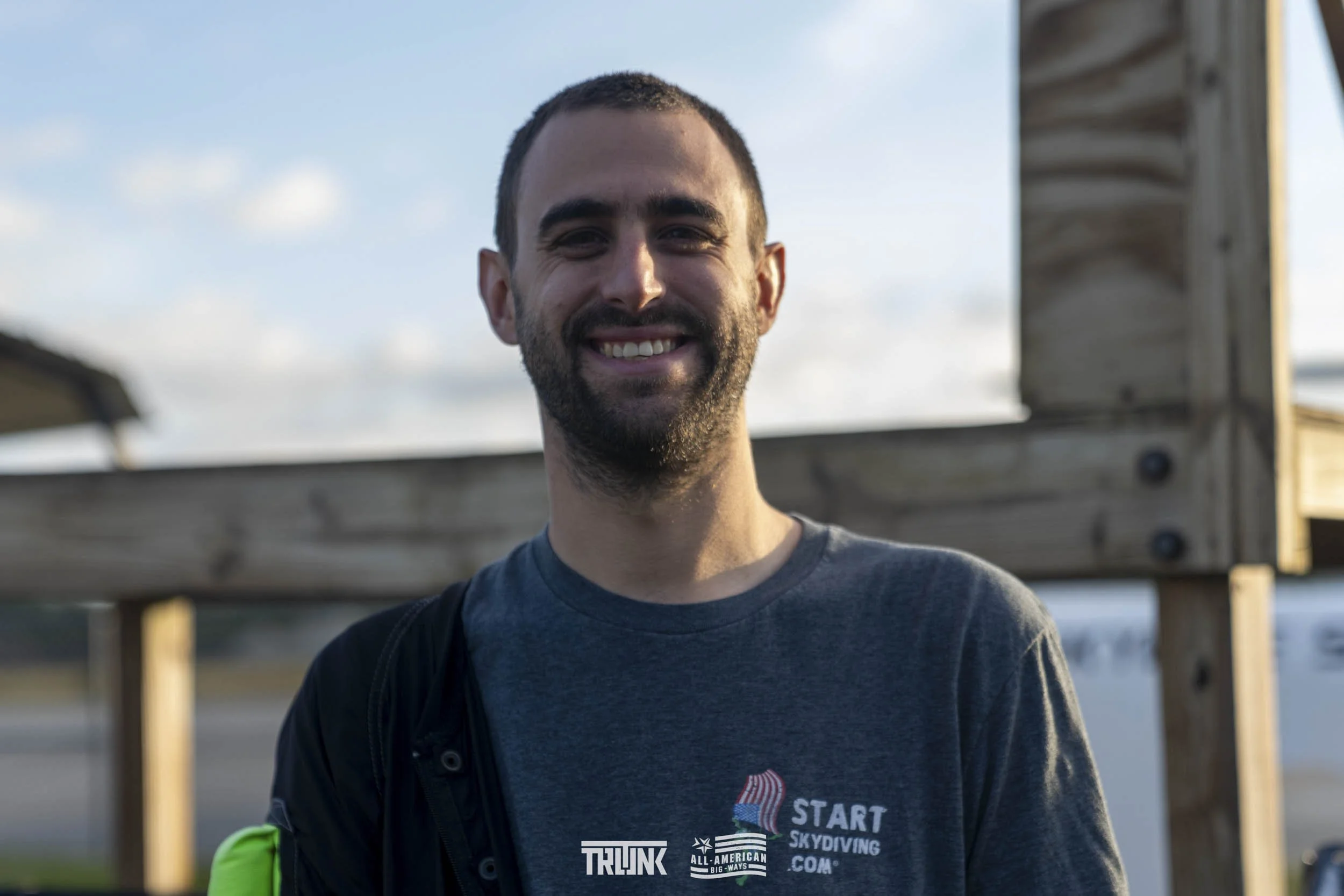A smiling man with dark hair and a beard standing outdoors near a wooden structure, wearing a dark grey t-shirt with a logo that says "START SKYDIVING.COM" and other texts.