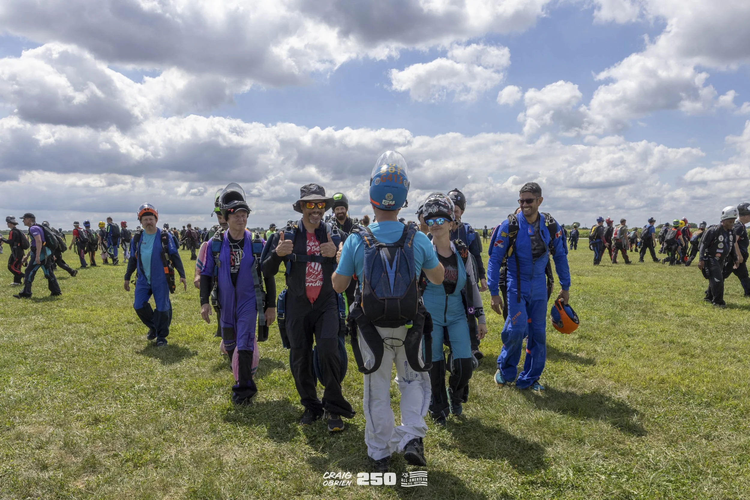 Group of skydivers walking on grassy field with open sky and clouds, some wearing helmets and jumpsuits, preparing for a jump.