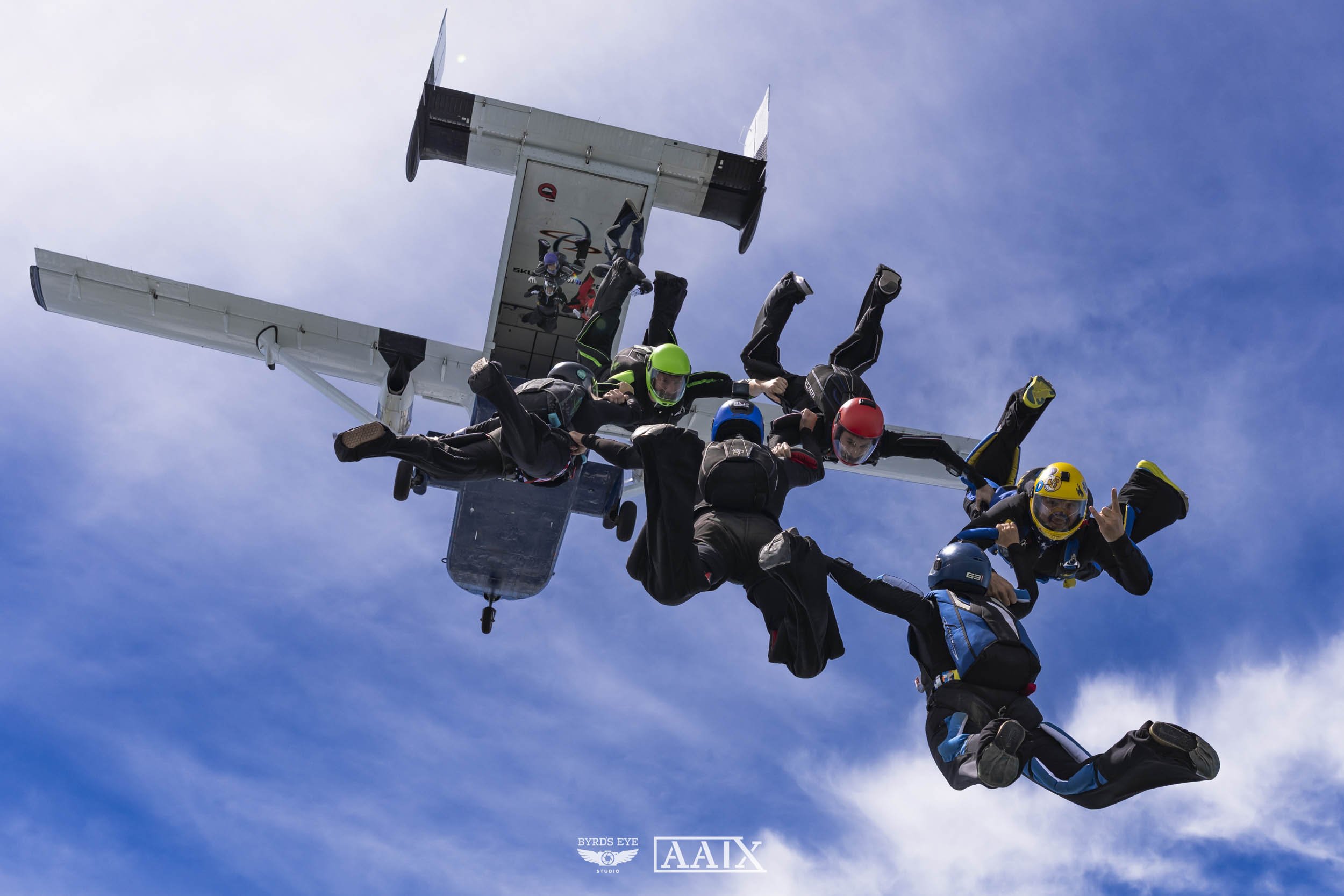 Skydivers holding hands in a formation in mid-air, with an aircraft flying overhead against a blue sky with wispy clouds.