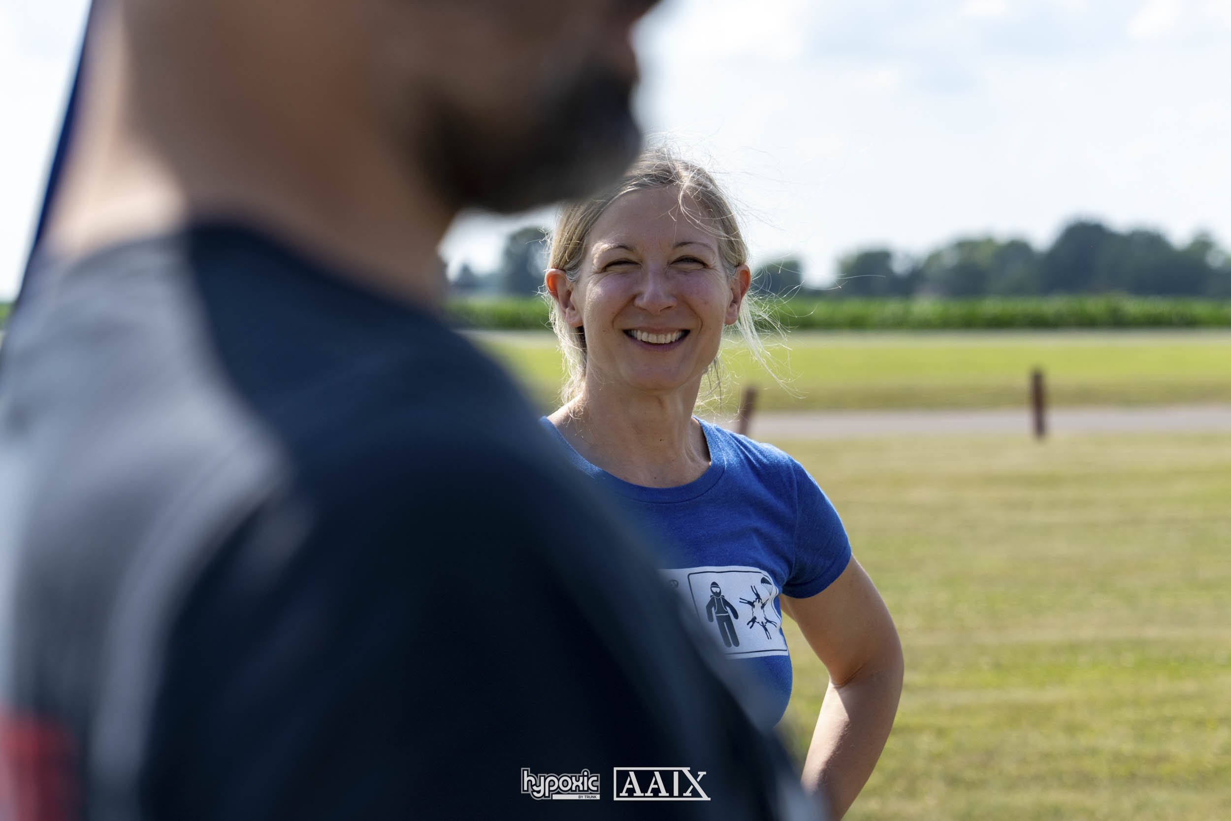 A woman smiling outdoors, with a person in the foreground whose face is out of focus, in a field or park on a sunny day.