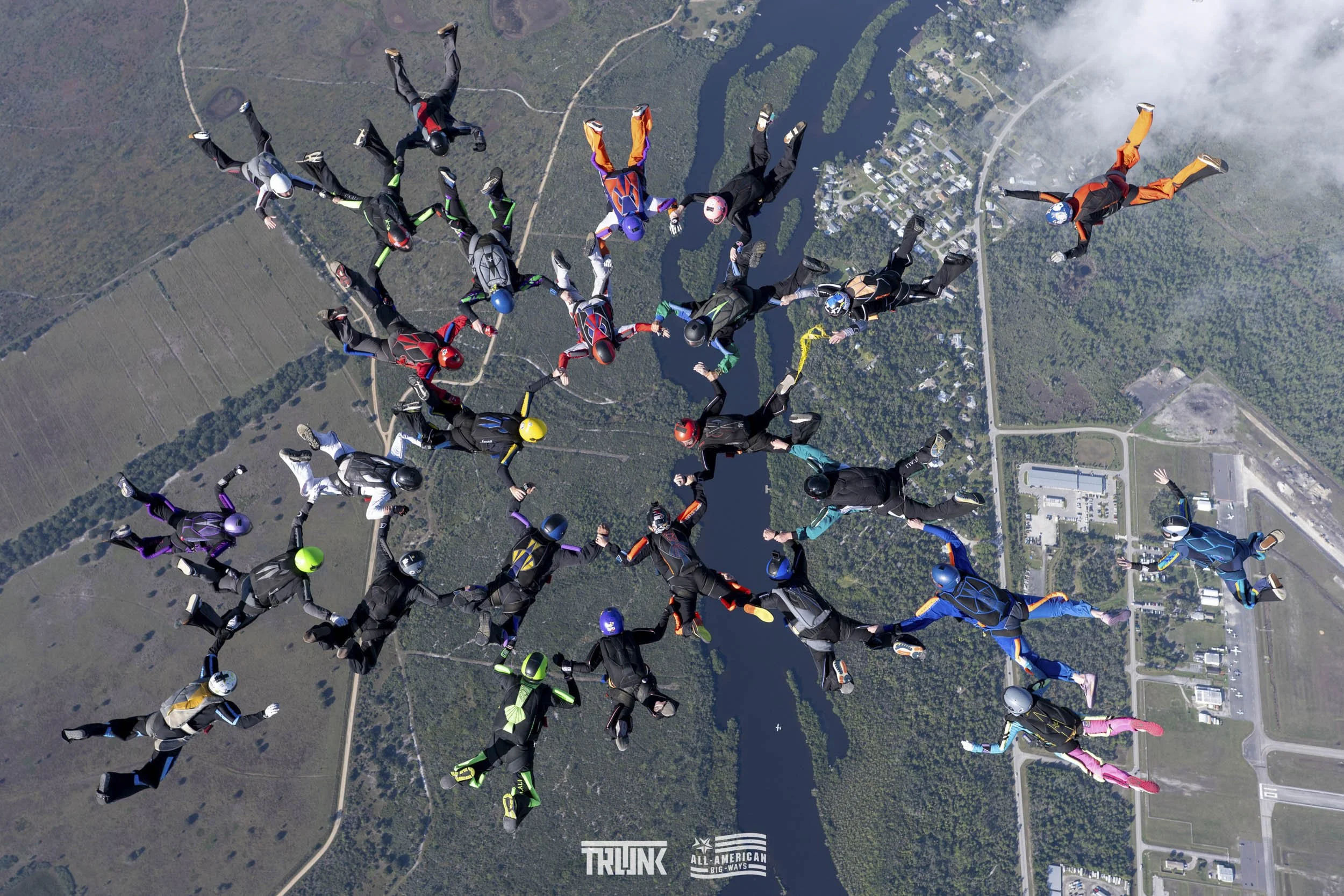 Group of skydivers holding hands while free-falling above a landscape with water, buildings, and trees.