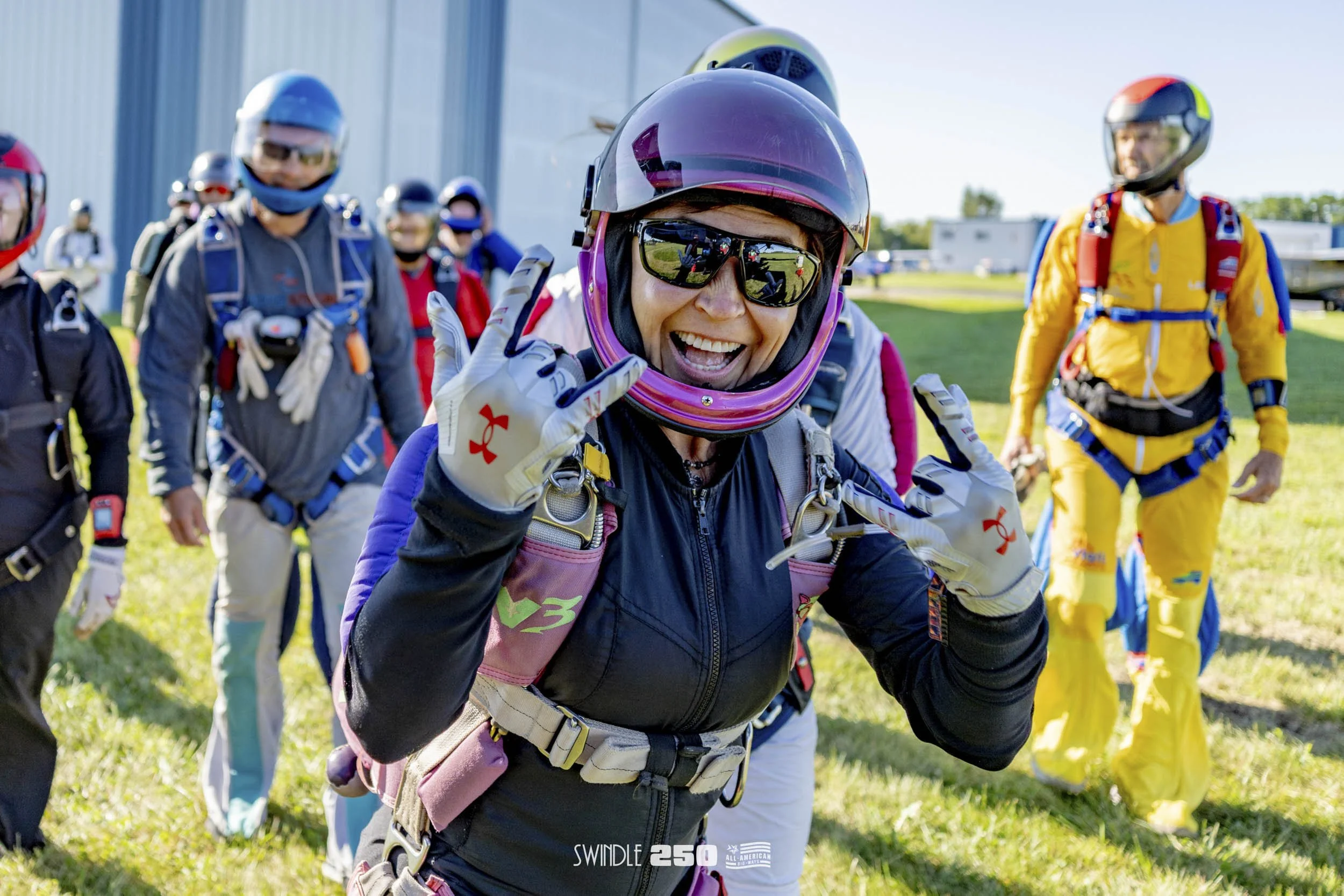 A group of skydivers standing on a grass field, with a woman in the foreground wearing a pink helmet, sunglasses, and a black jumpsuit, smiling and making hand gestures, while others in colorful jumpsuits stand behind her.