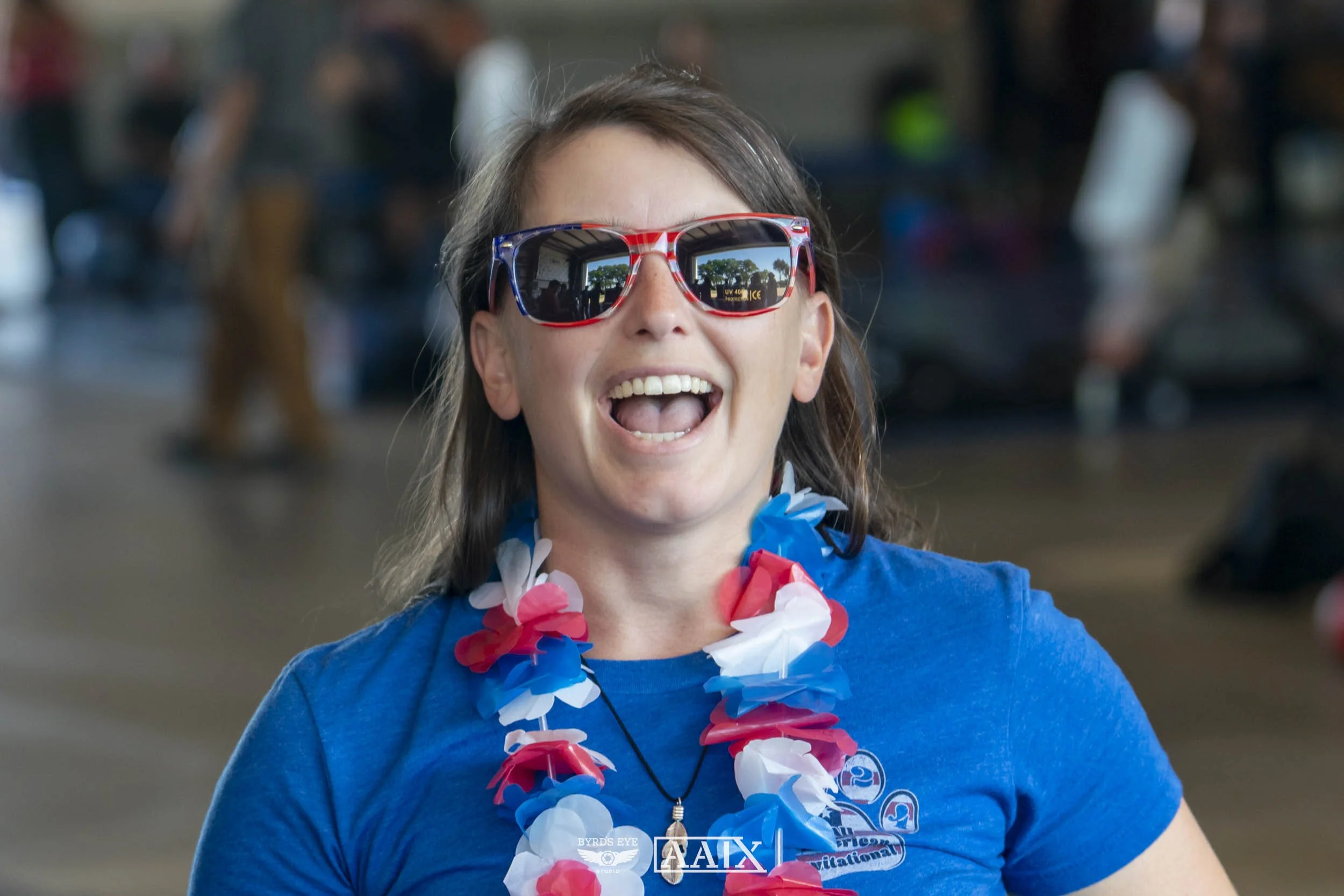 A woman at an airport or outdoor event wearing American flag themed sunglasses, a blue shirt, and a red, white, and blue leis, smiling with an open mouth.