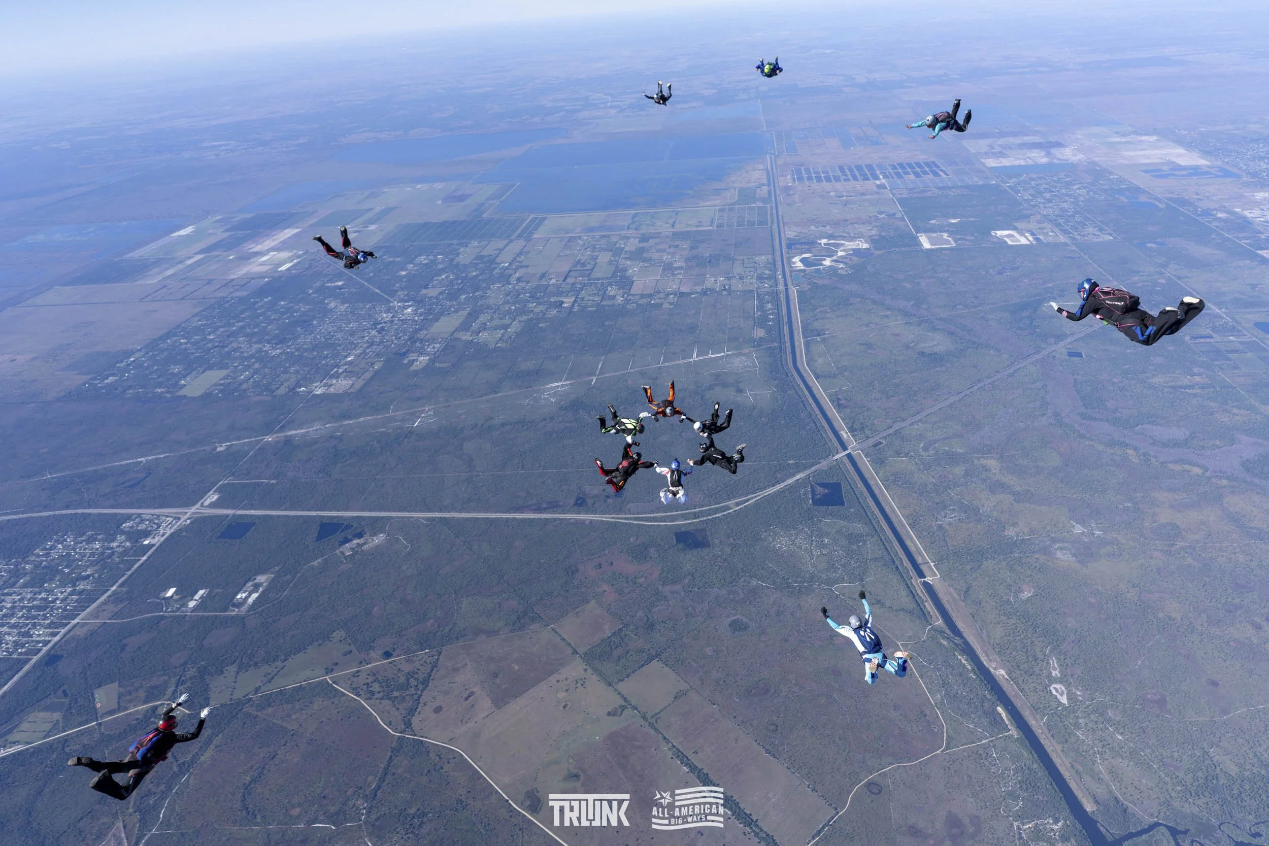 Skydivers in free fall forming a circle over a landscape of farmland and highways, with some skydivers scattered around the circle.
