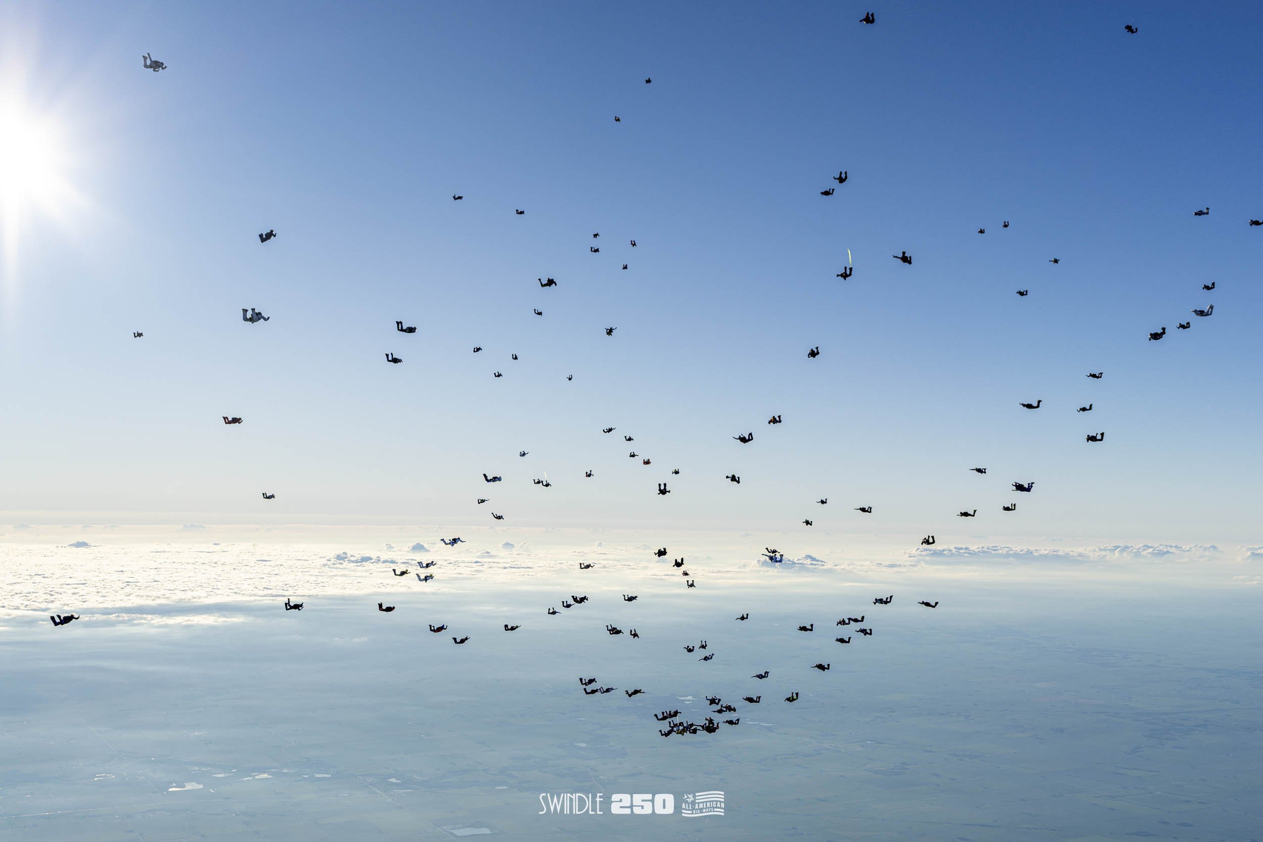 A large number of skydivers in black jumpsuits with parachutes are descending over a snow-covered landscape on a clear day with a bright sun.