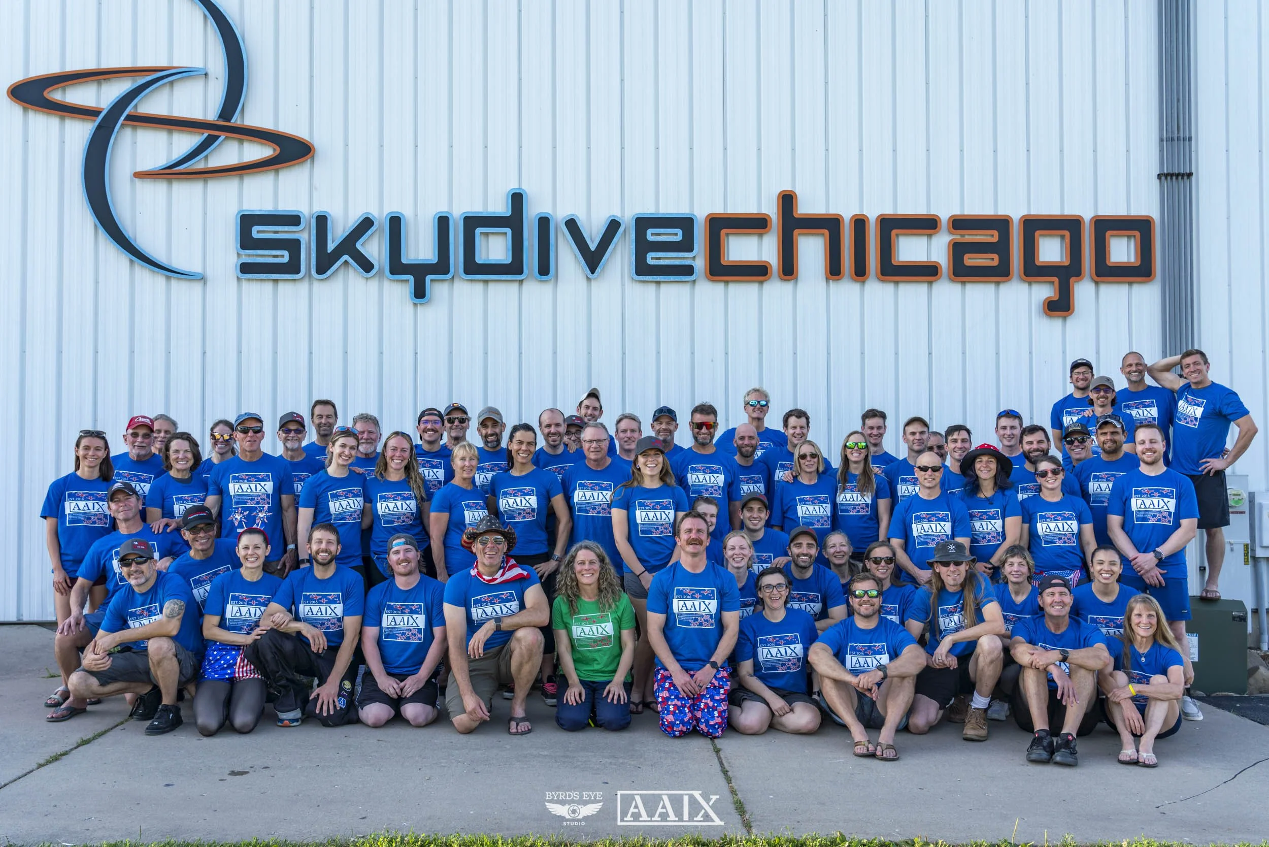 A large group of people wearing matching blue T-shirts with the AAIX logo, posing in front of a building with a neon sign that reads Skydiving Chicago. The group is smiling and standing on a concrete surface outside the skydiving facility.
