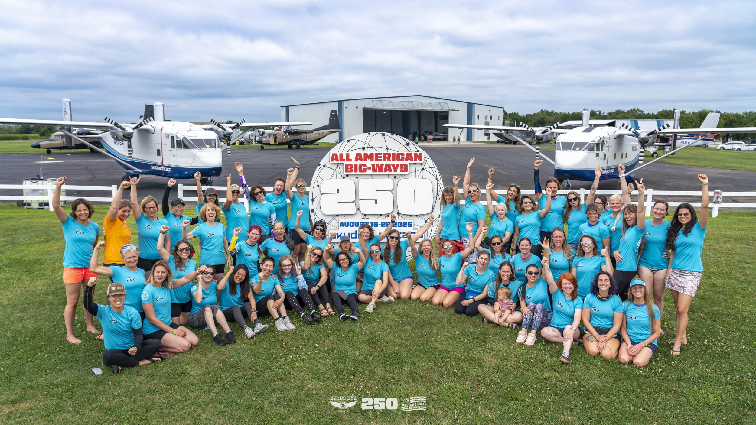 Group of women celebrating in front of a sign reading 'All American Big-Ways 250' at an airfield with several small airplanes in the background.
