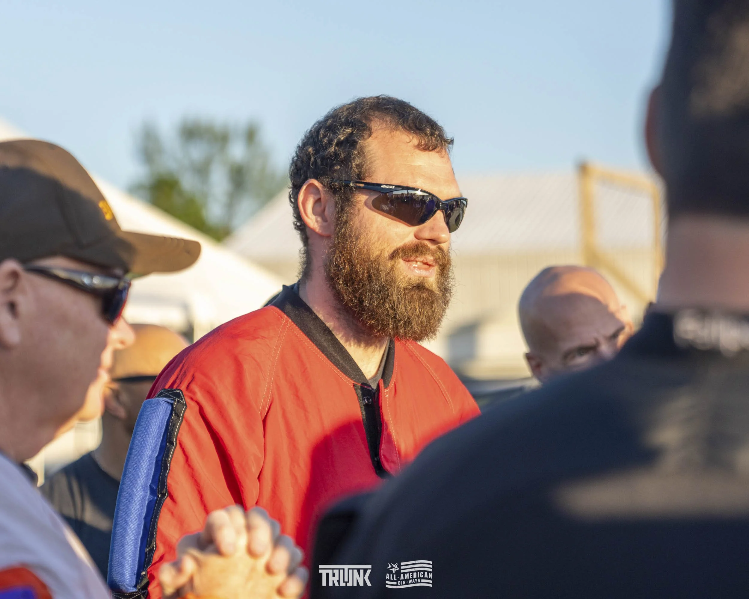 A man with a full beard and sunglasses wearing a red jacket is in a group conversation outdoors during daytime, with people around him and tents in the background.