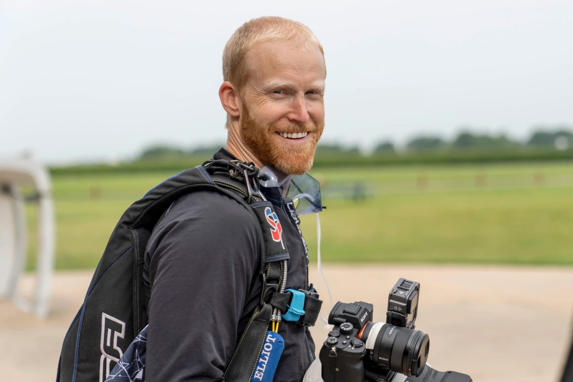 A man with red hair and beard smiling while holding a camera, outdoors in a grassy field with a cloudy sky.