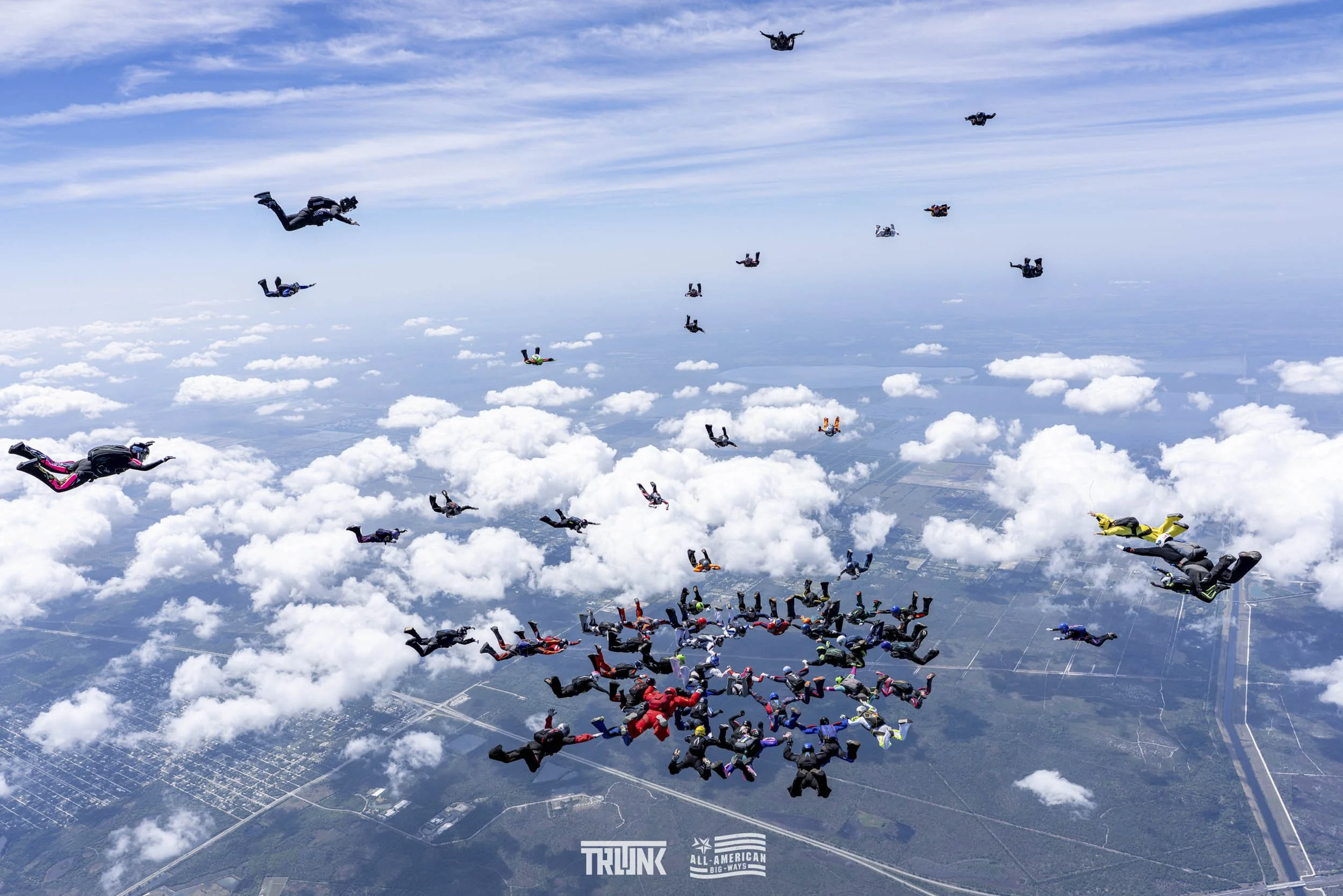 Skydivers in free fall over a landscape with clouds below and a blue sky, forming a circle pattern.