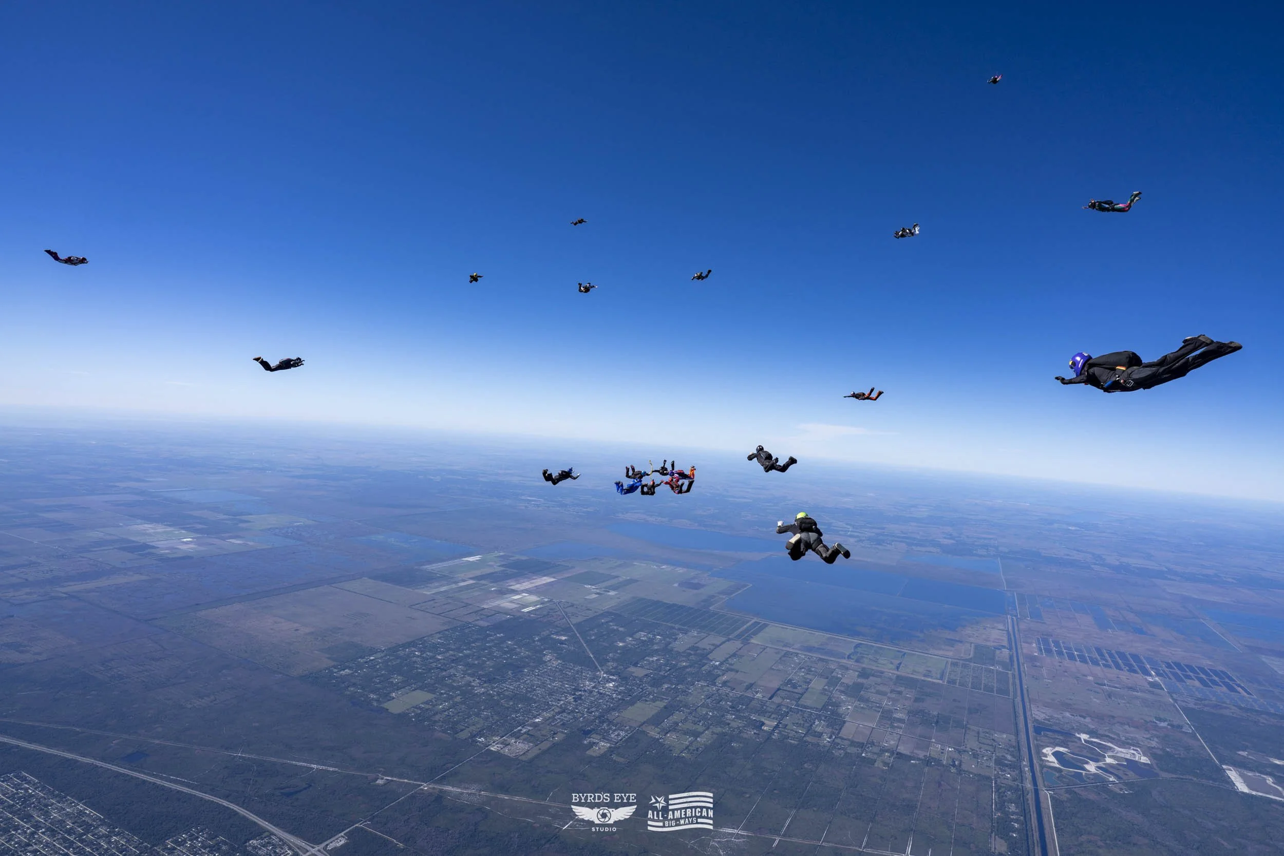 Group of skydivers in freefall over a patchwork farmland landscape under a blue sky.