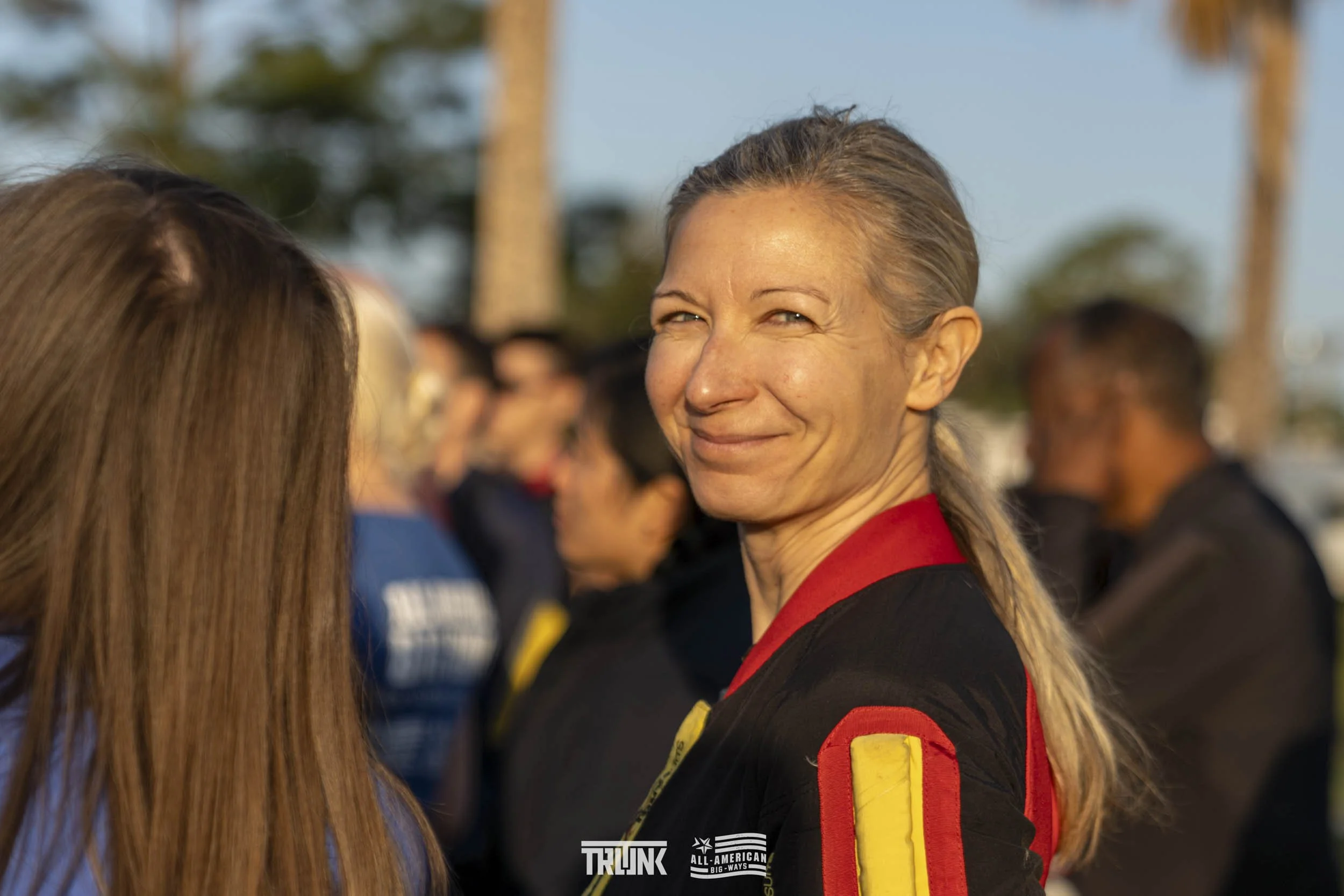 A woman with gray hair tied back, smiling and looking towards the camera, wearing a black and red uniform with yellow patches, outdoors among other people and palm trees.