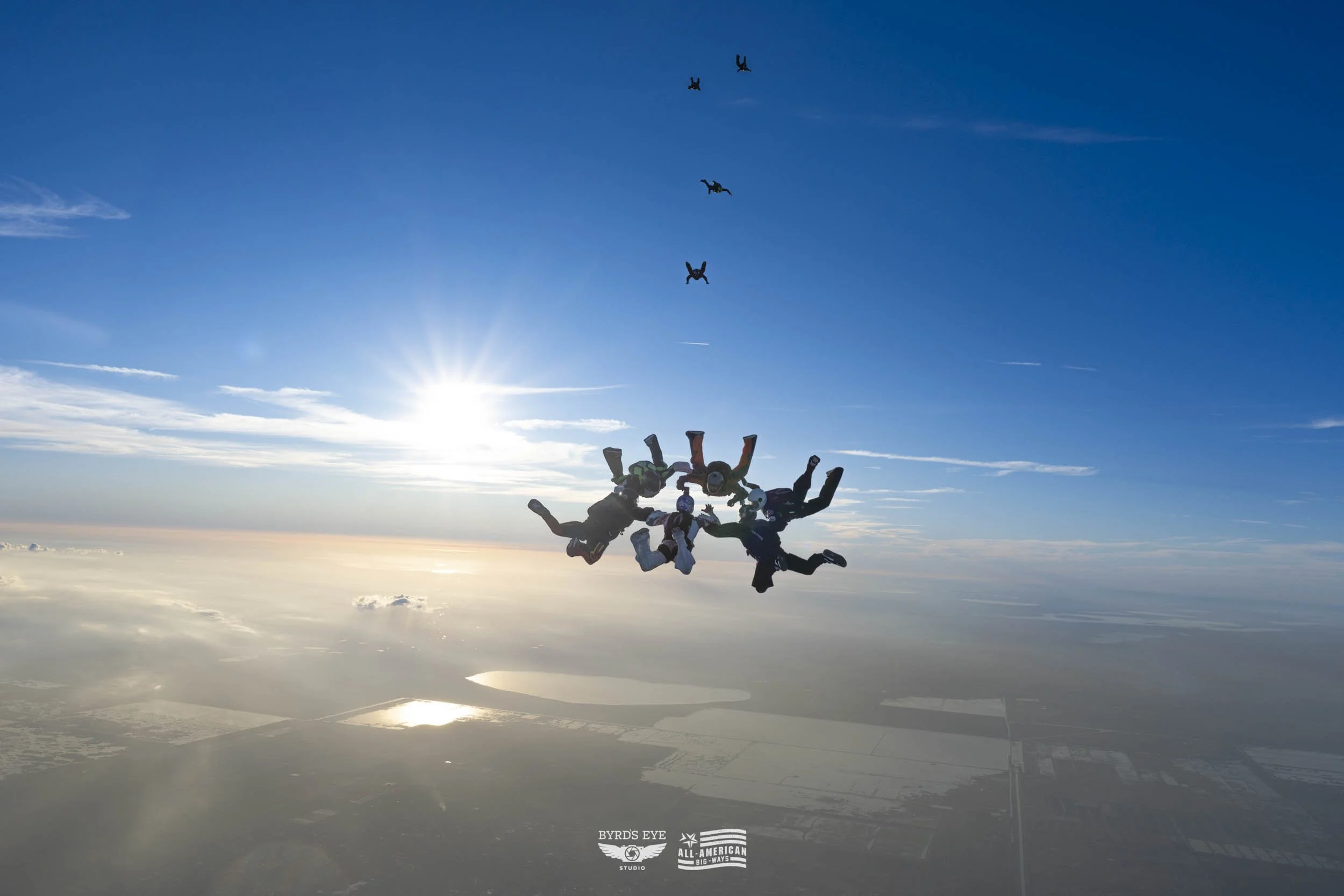 Group of skydivers in mid-air formation, with a clear blue sky and the sun low on the horizon, over landscape and water below.