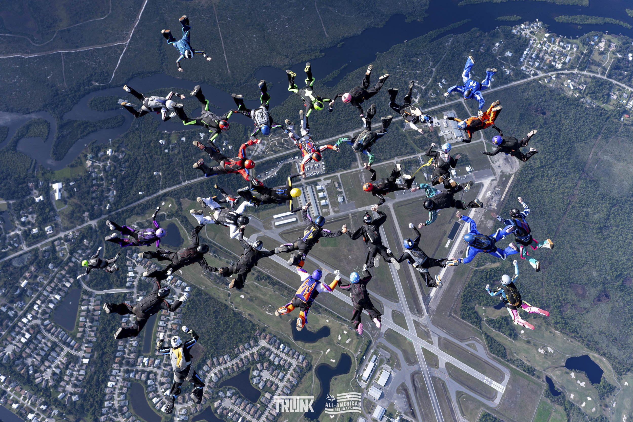Group of skydivers in free fall over a landscape with lakes, forests, and buildings.