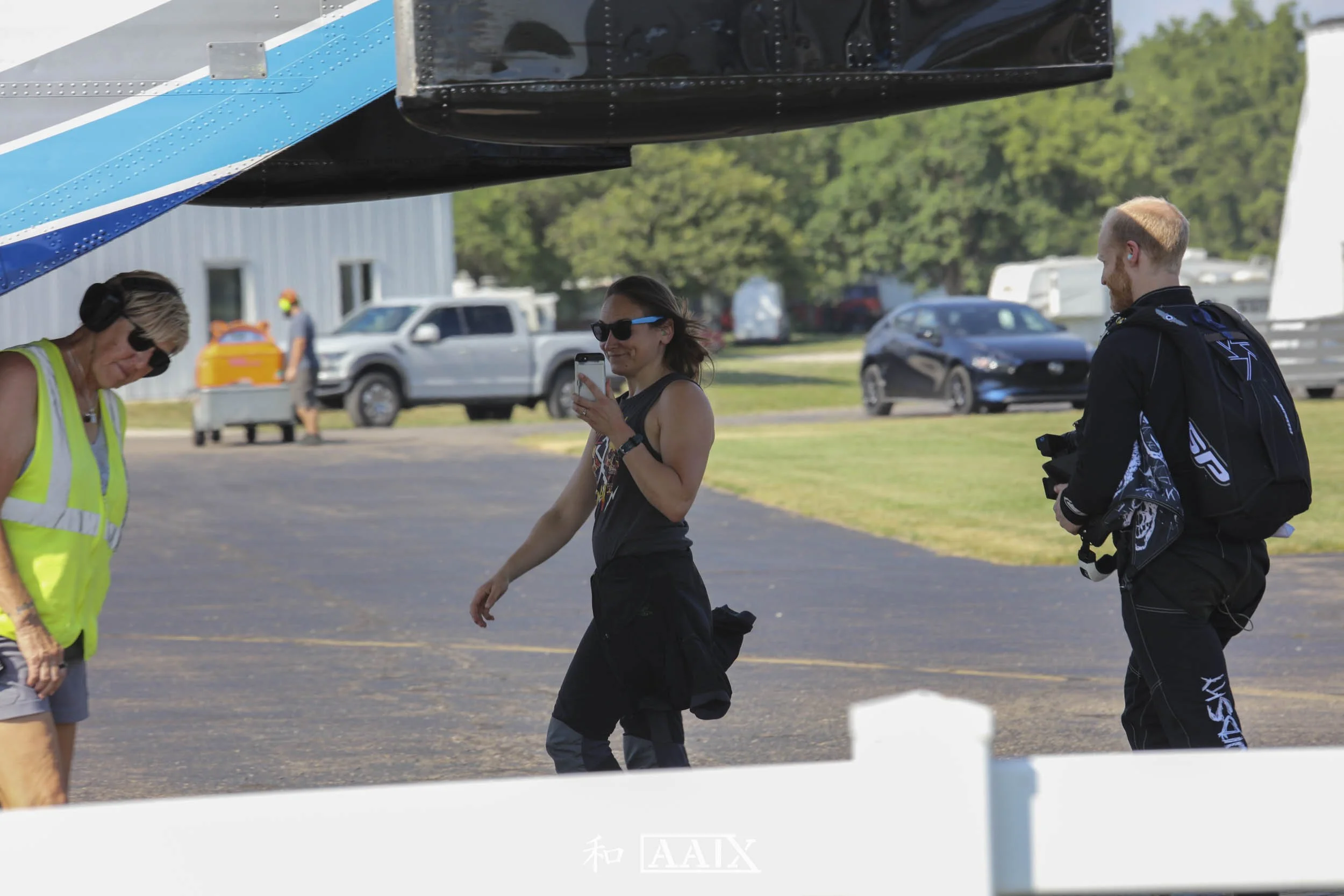 Three people standing outdoors near an aircraft, with one woman taking a photo or selfie; a woman in a yellow safety vest, a woman with sunglasses and a black tank top, and a man with a backpack holding a camera, with cars and trees in the background