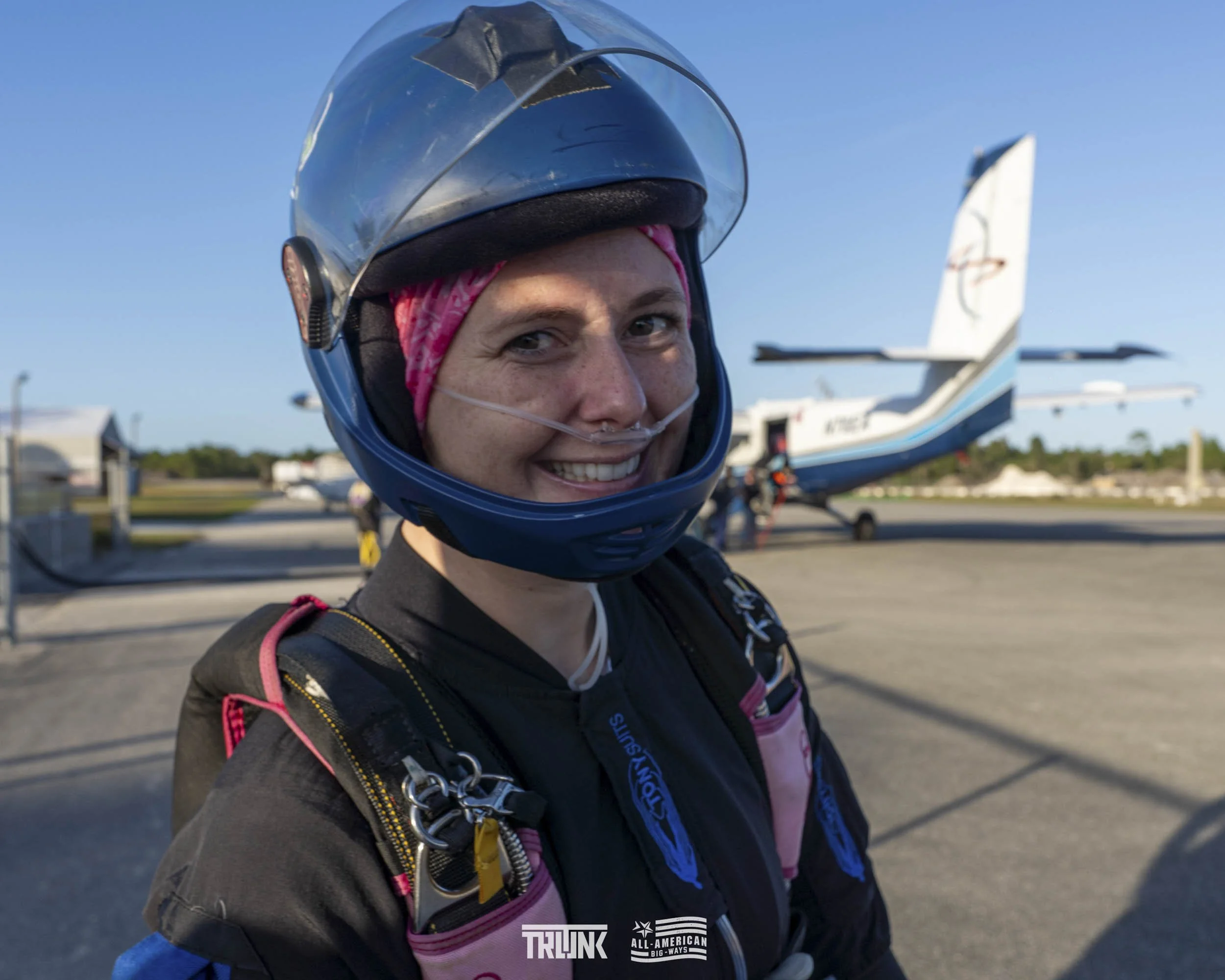 A woman in skydiving gear, wearing a helmet and pink headband, smiling at the camera with an airplane in the background on a clear day.