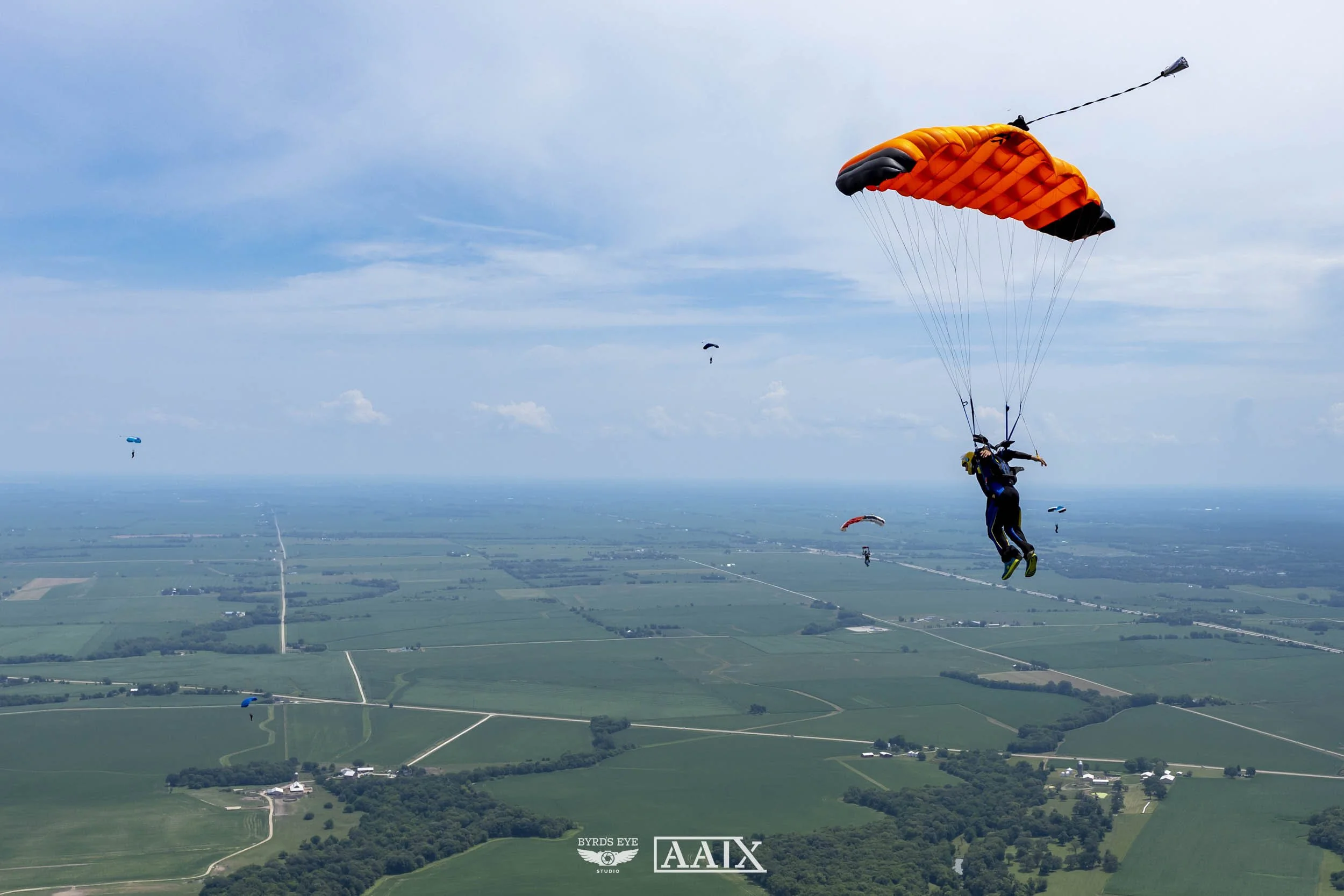 Skydivers descending with parachutes over a rural landscape with fields and roads.