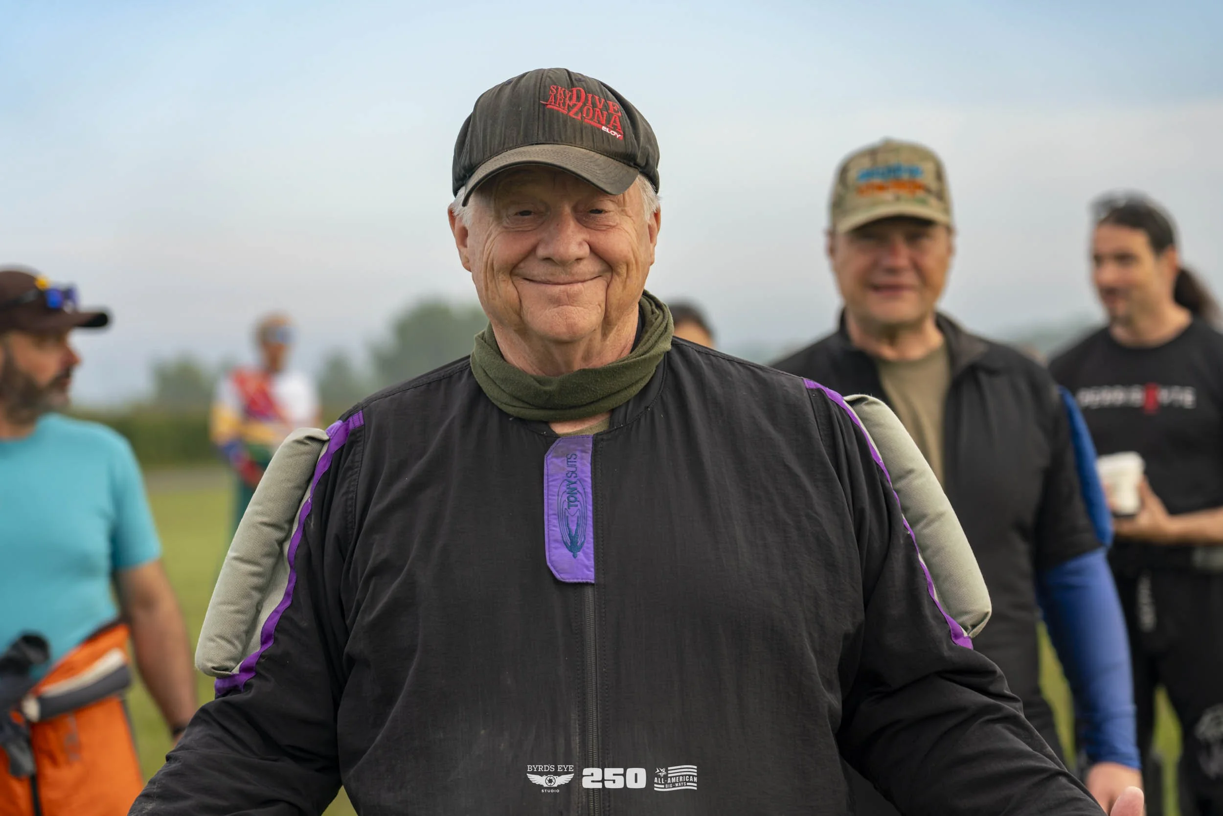 An elderly man smiling, wearing a black cap with red text, a black jacket with purple accents, and a green neck gaiter, standing outdoors with a group of people in the background.