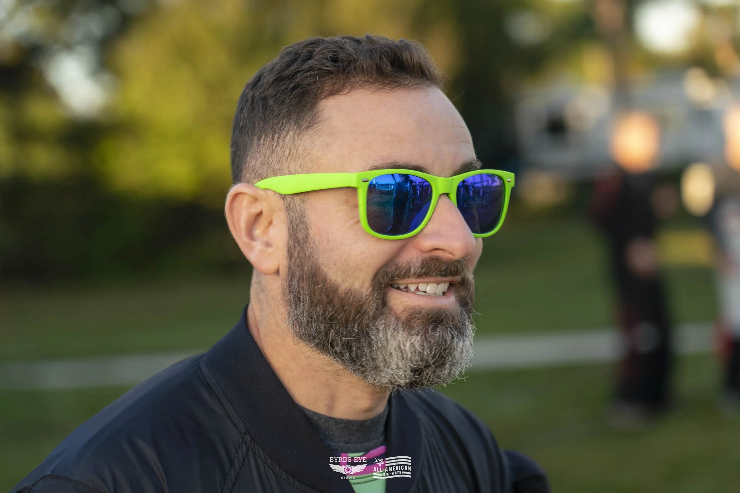 Close-up of a smiling man with a beard wearing brightly colored green sunglasses, outside with trees in the background.