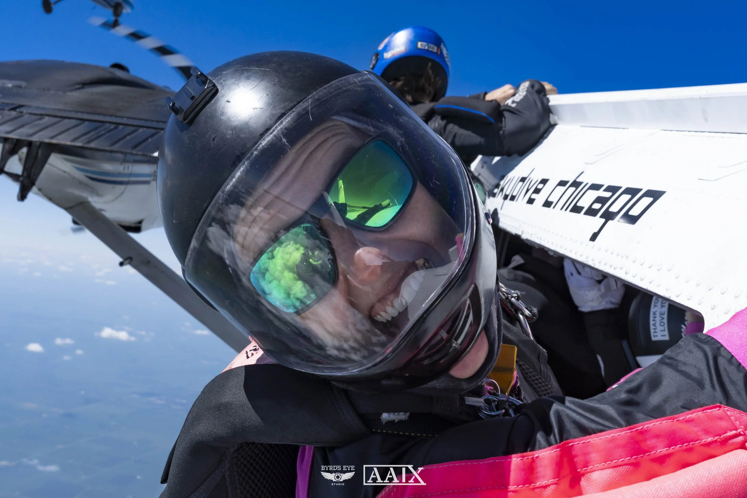 A skydiver taking a selfie after jumping from an aircraft, with another skydiver lying in the plane in the background. The sky is clear and blue.