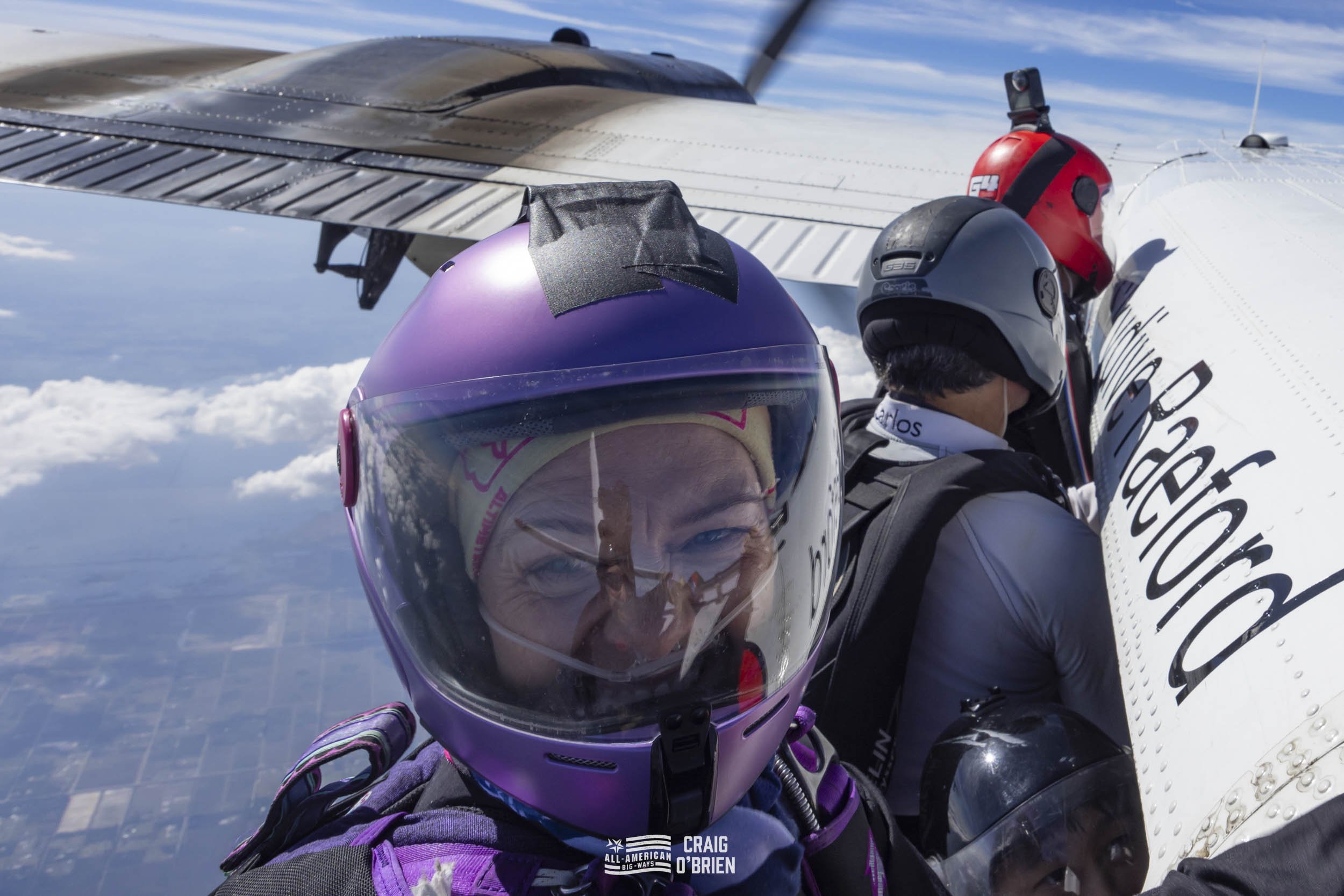 Two skydivers in helmets and jumpsuits clinging to the side of an aircraft in mid-flight, with a clear blue sky and clouds below.