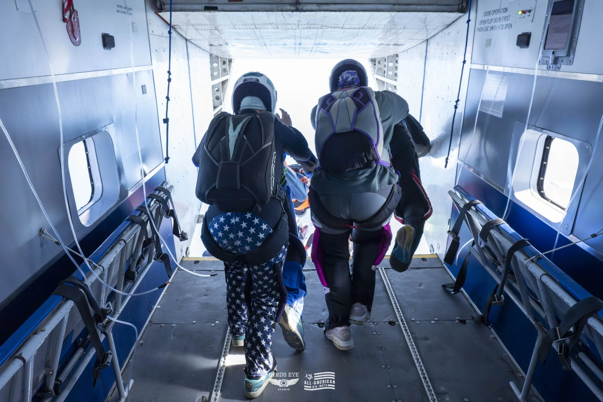 Three skydivers preparing to exit an aircraft, seen from behind, with parachuting gear and helmets.
