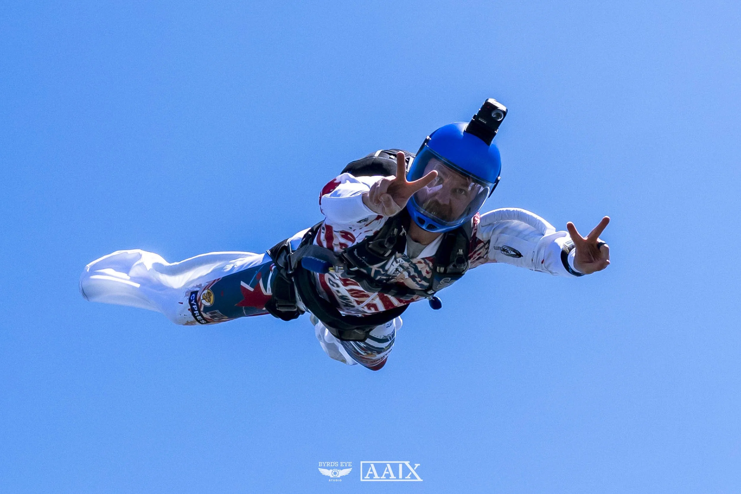 A skydiver in a white jumpsuit with red and black accents, wearing a blue helmet with a camera mounted on top, is free-falling against a clear blue sky, making peace signs with both hands.