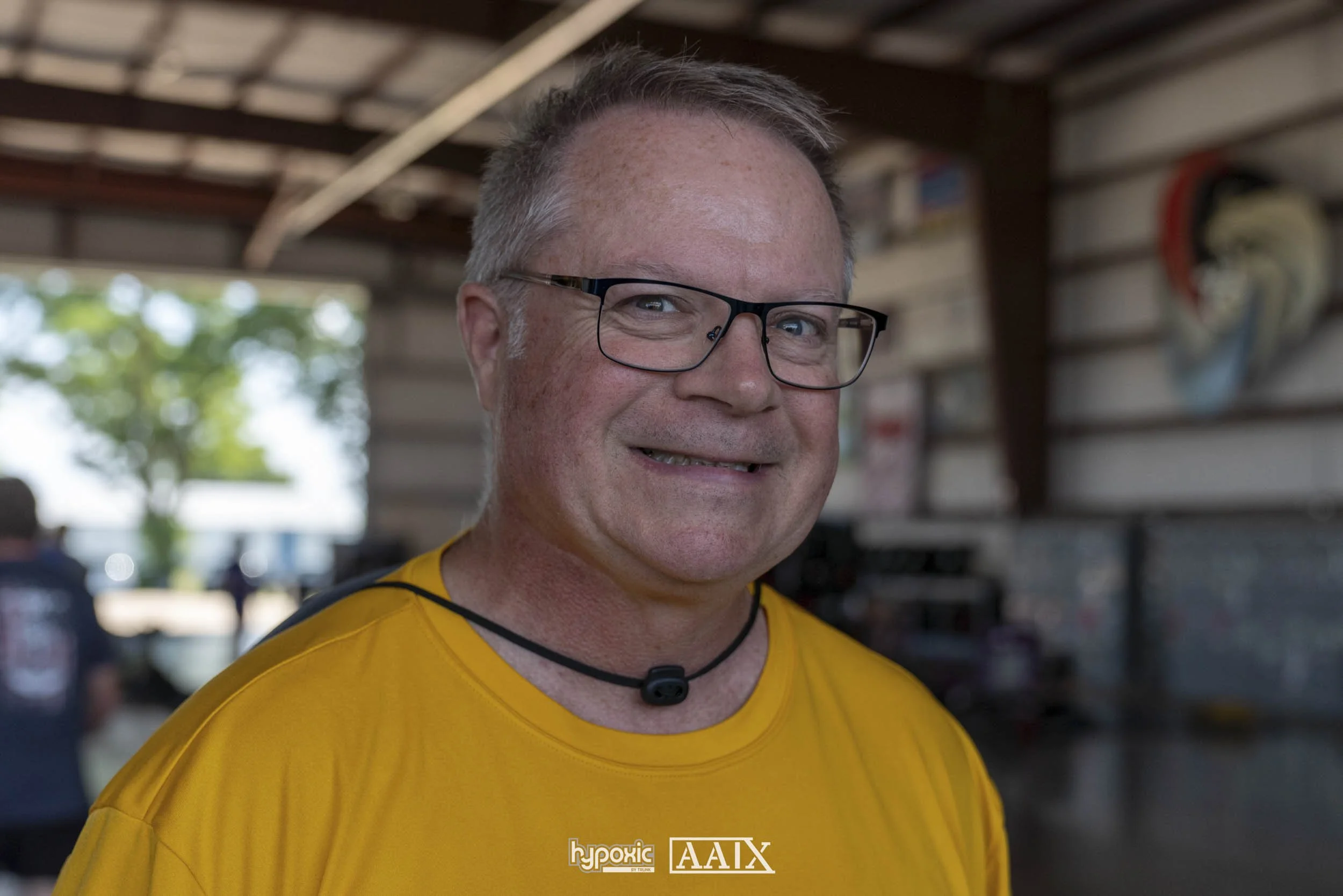 A middle-aged man with short gray hair, glasses, and light skin, smiling while wearing a yellow shirt and a black necklace. He is inside a large barn or warehouse with other people and trees visible outside in the background.
