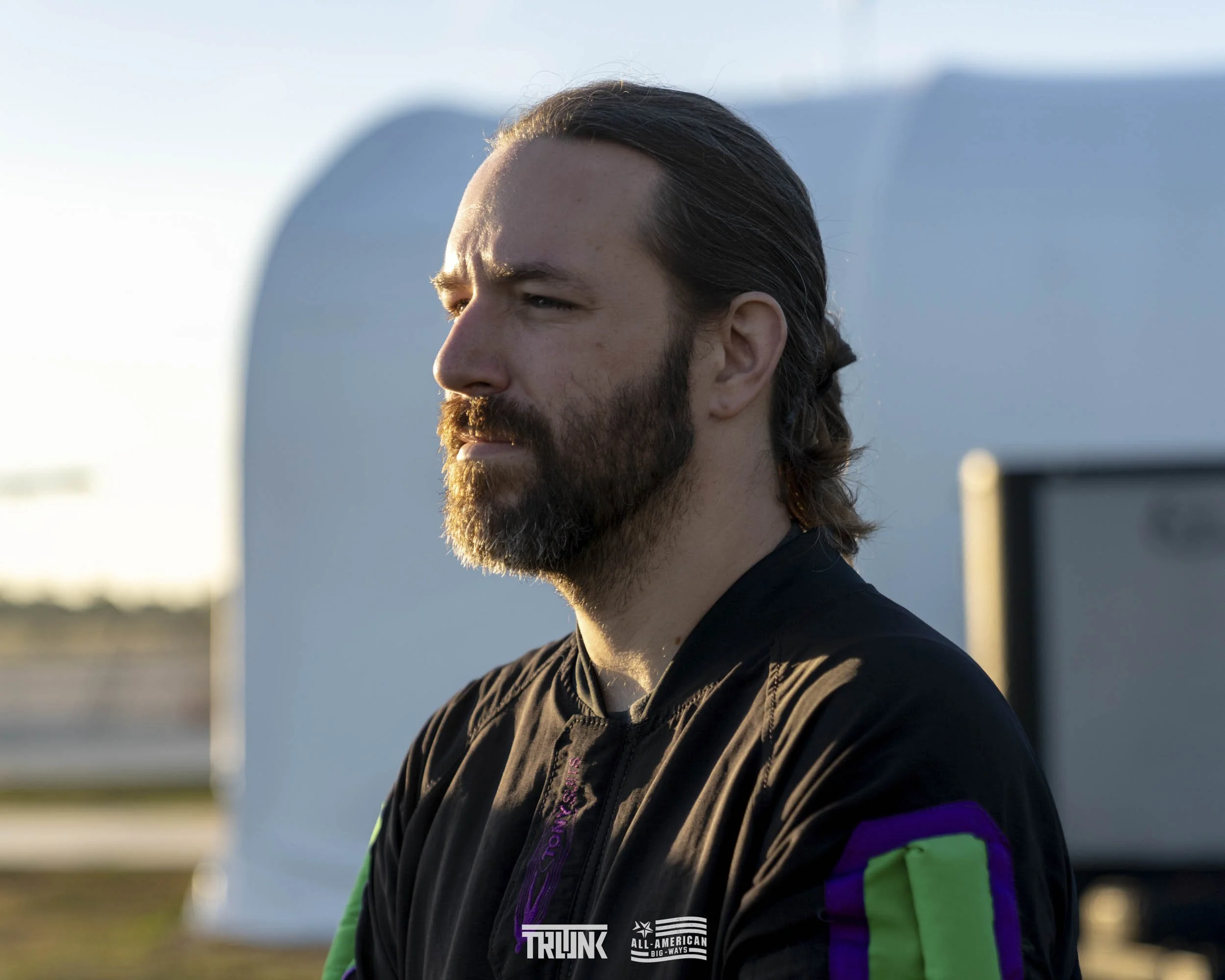A man with long brown hair and a beard, looking to the left, outdoors in natural light, wearing a black jacket with purple and green accents, with a large white trailer in the background.