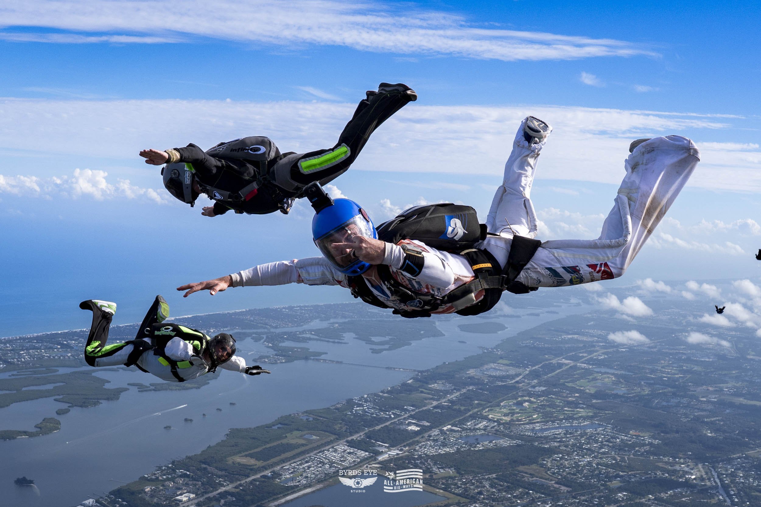 Three skydivers in free fall above a landscape with rivers and fields, wearing helmets, jumpsuits, and harnesses.