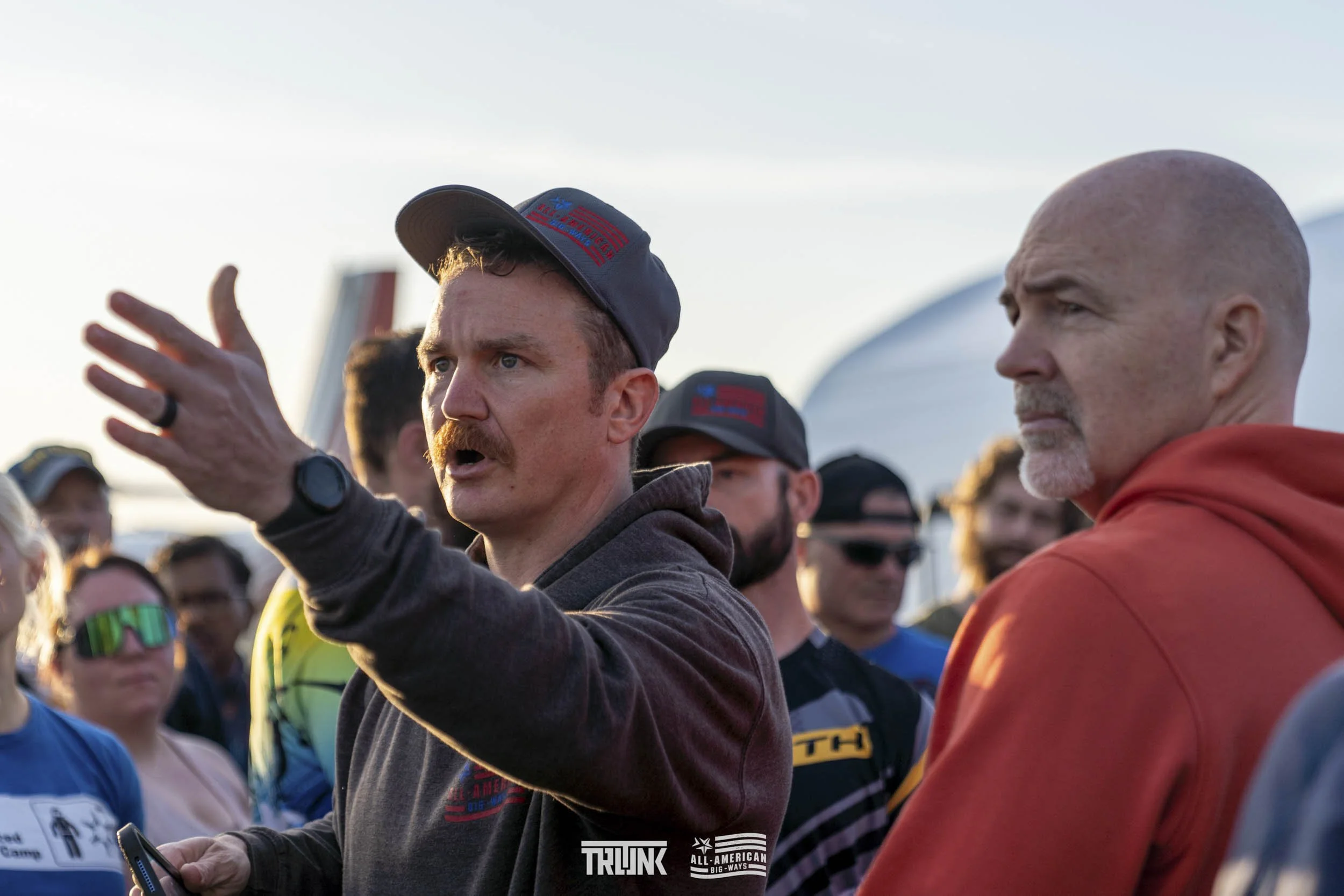 A man with a mustache and wearing a baseball cap is speaking passionately, gesturing with his hand at a crowd of people outdoors during daytime.