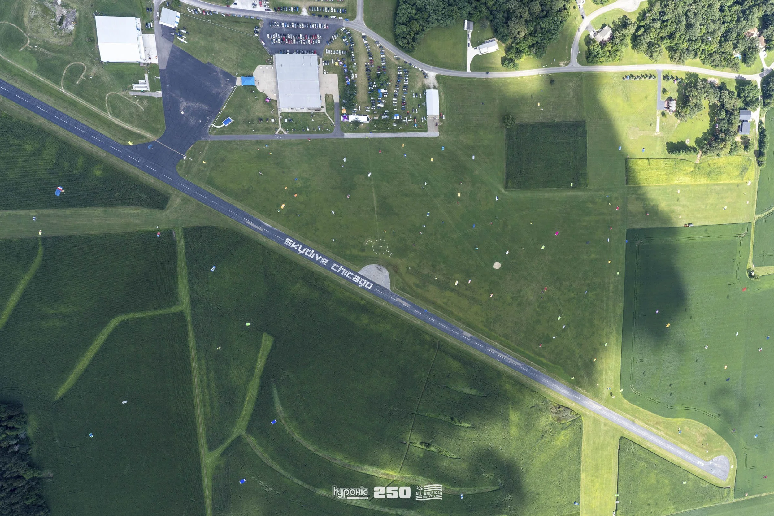 Aerial view of a park with a large, open grassy area, marked with numerous colorful balloons. There are trees, roads, and buildings nearby, including a parking lot. The road has the words 'Welcome to Chicago' painted on it.