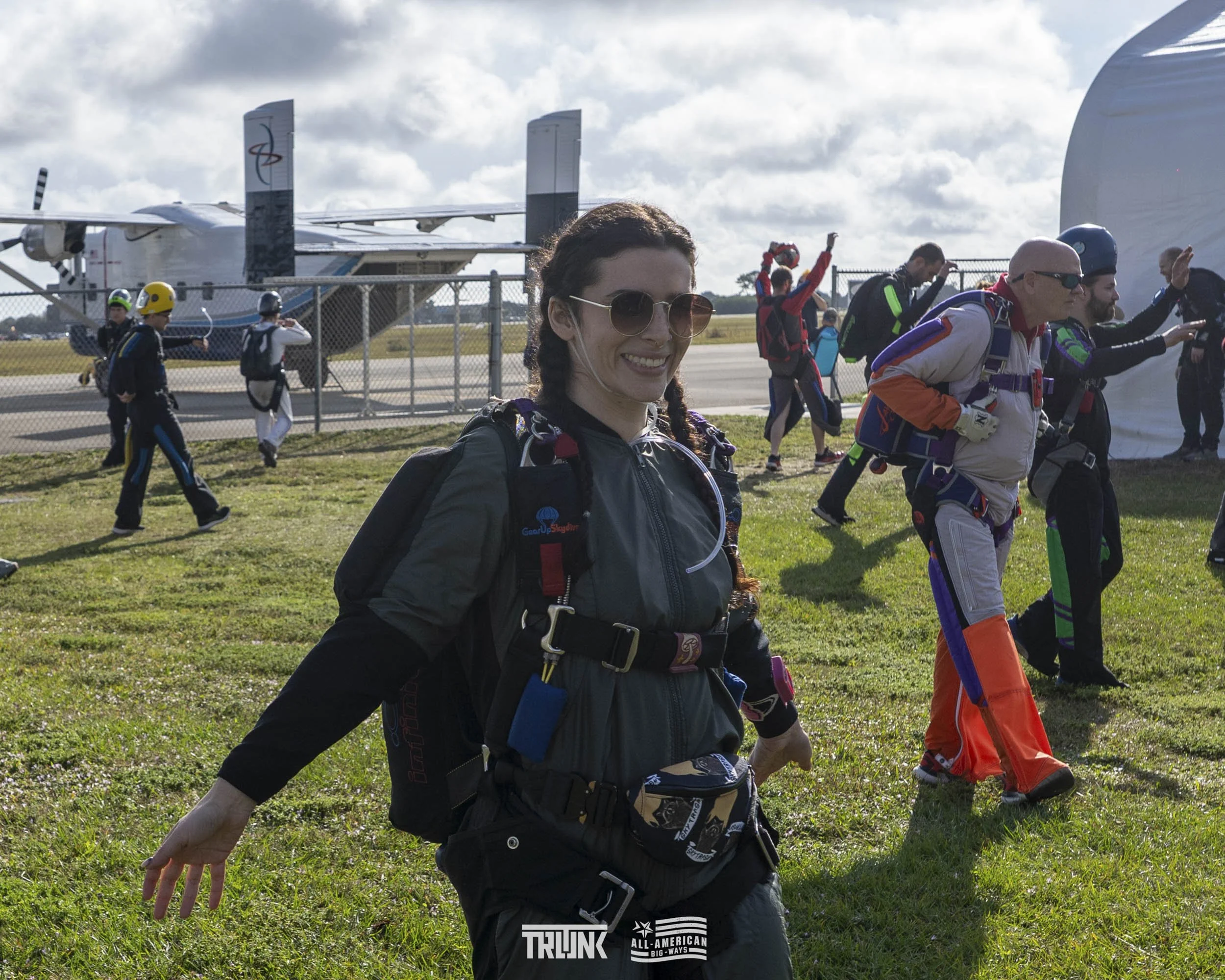 A woman smiling and wearing skydiving gear, standing on grass with a group of skydivers and an aircraft in the background at an airfield.