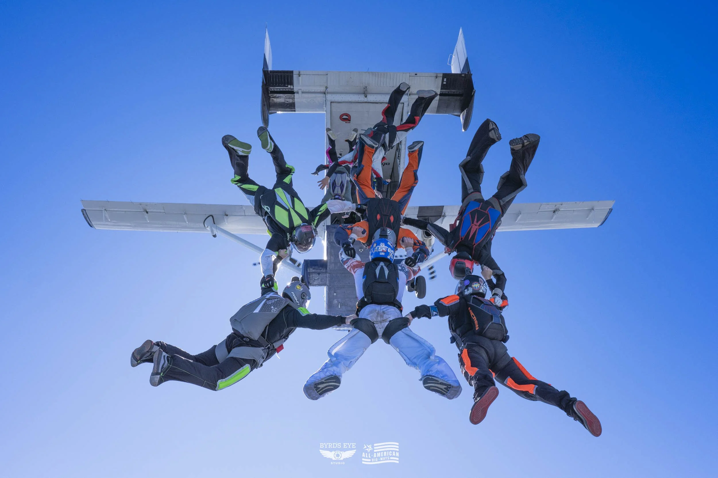 Skydivers forming a circle holding hands while falling towards the ground, with an airplane in the background against a clear blue sky.