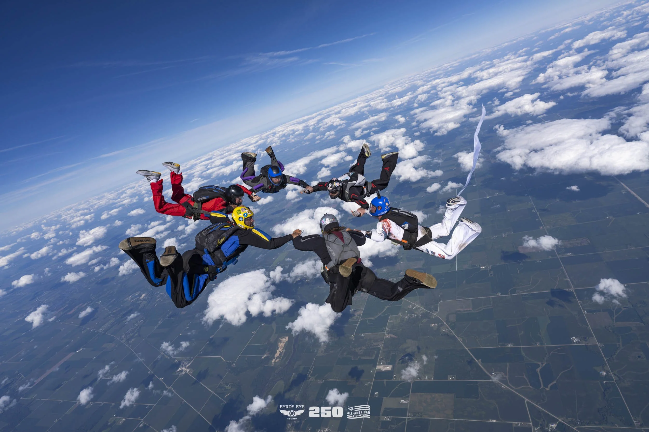 Skydivers holding hands in a circle mid-air above the clouds, with a clear blue sky and farmland below.