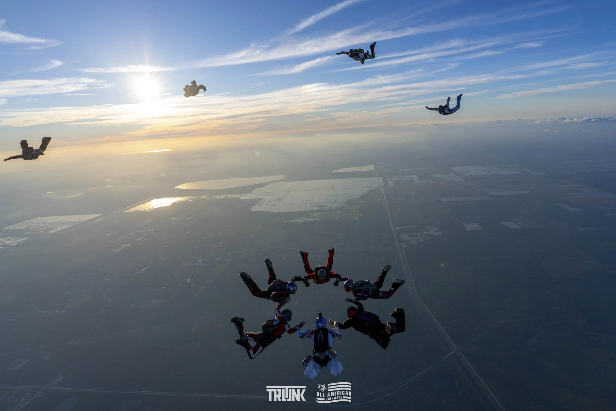 Skydivers freefalling in a group over a landscape with lakes and fields, sunset sky with clouds.