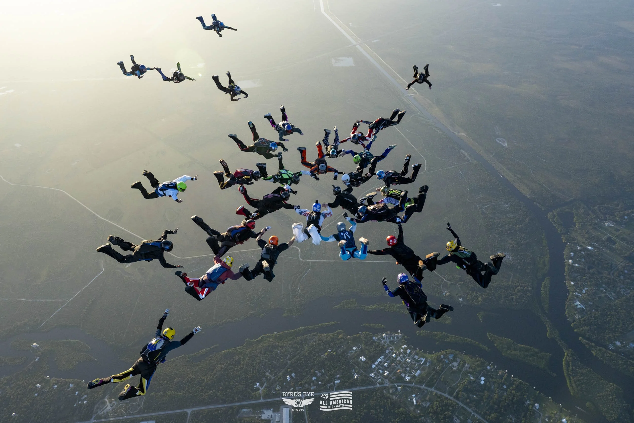 Group of skydivers in free fall over a landscape, wearing colorful jumpsuits and helmets.