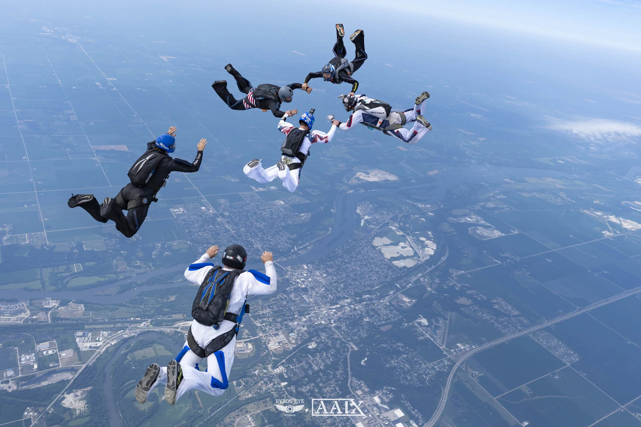 Group of skydivers holding hands in formation during free fall over a landscape with water and roads.