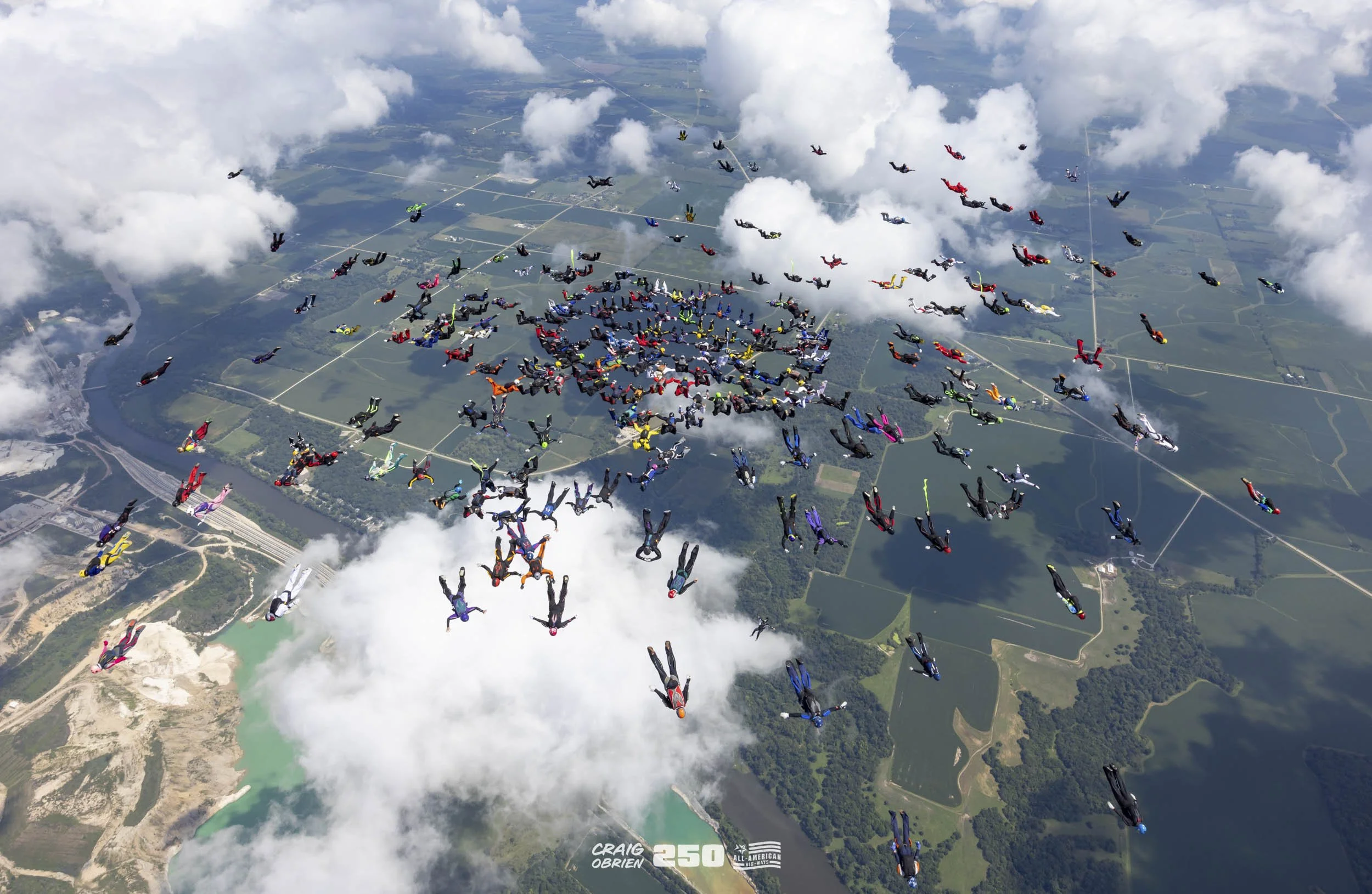 A large group of skydivers in colorful suits free-falling from the sky over green landscape, with some clouds at altitude.