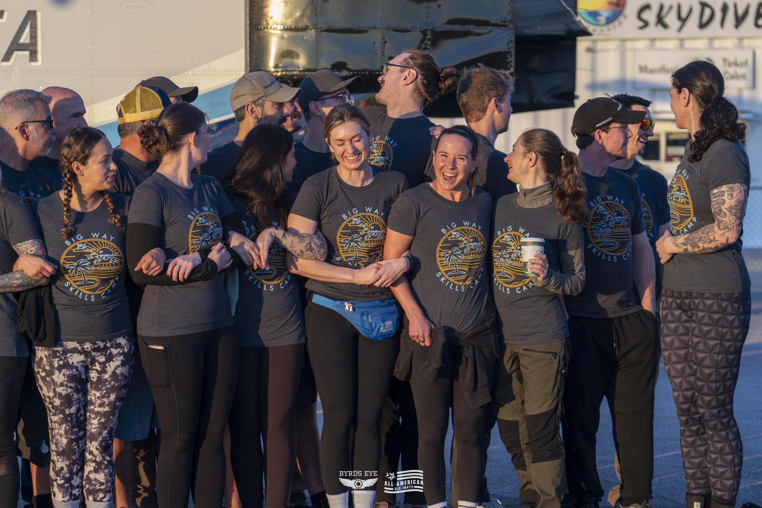 A group of people wearing matching dark gray shirts with a logo, standing closely together outdoors, smiling and chatting during daylight.