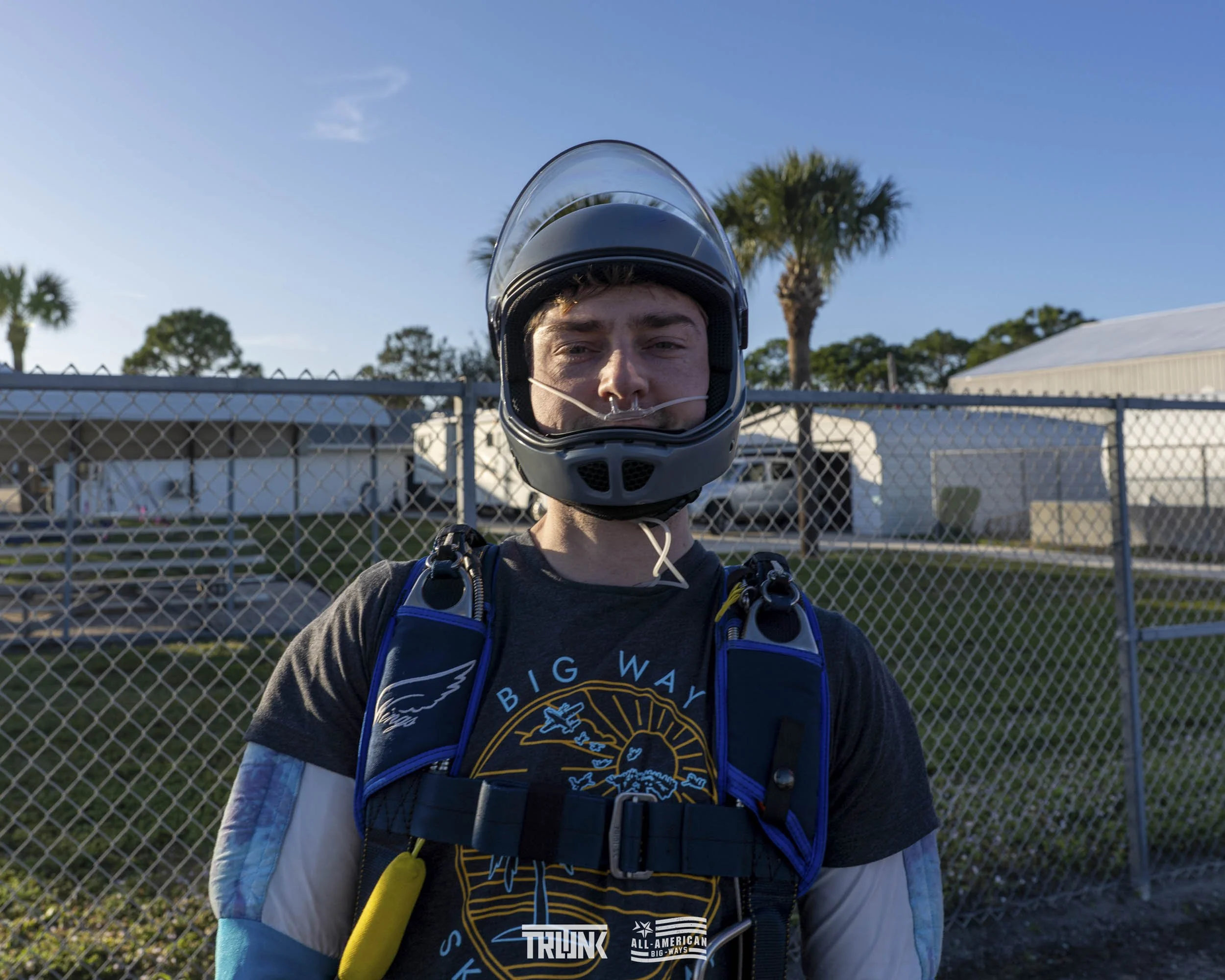 A young man wearing a motorcycle helmet and motorcycle gear standing outdoors in front of a chain-link fence with palm trees and a blue sky in the background.