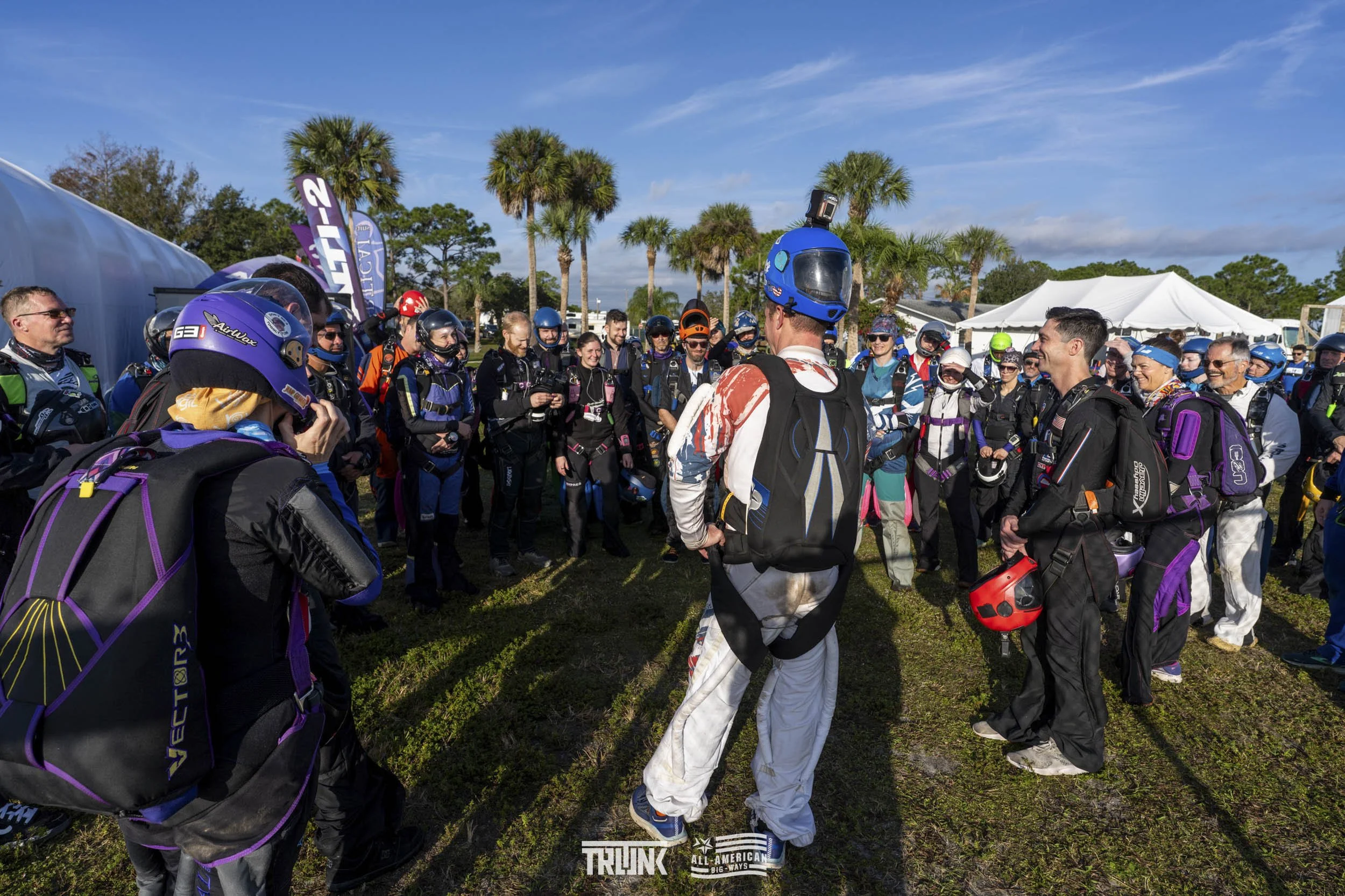 Group of skydivers gathered outdoors on a sunny day near palm trees, listening to an instructor in a jumpsuit, with some in helmets and parachutes visible.