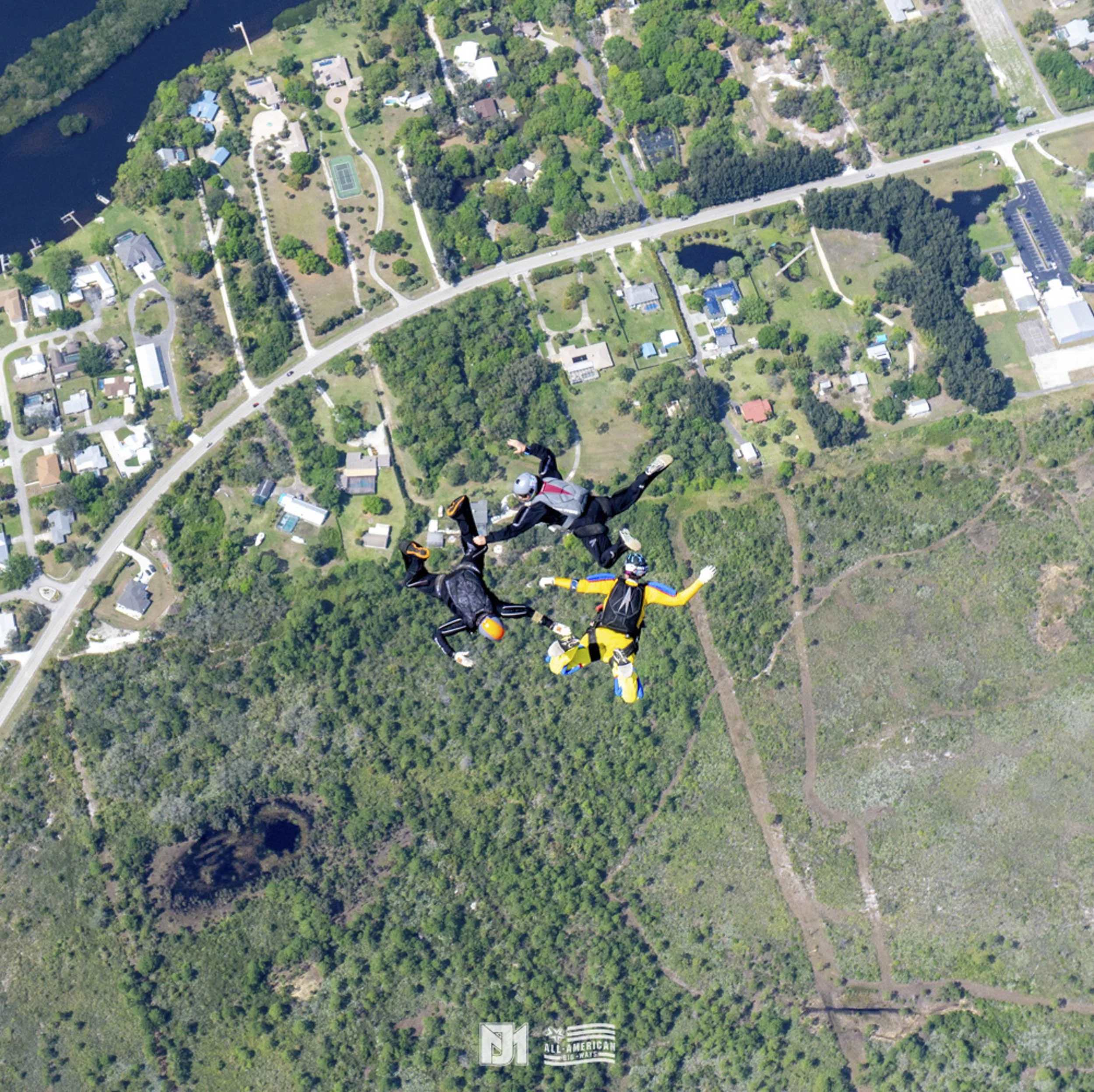 Three skydivers in freefall above a green, wooded landscape with a residential area, a lake, and roads visible below.