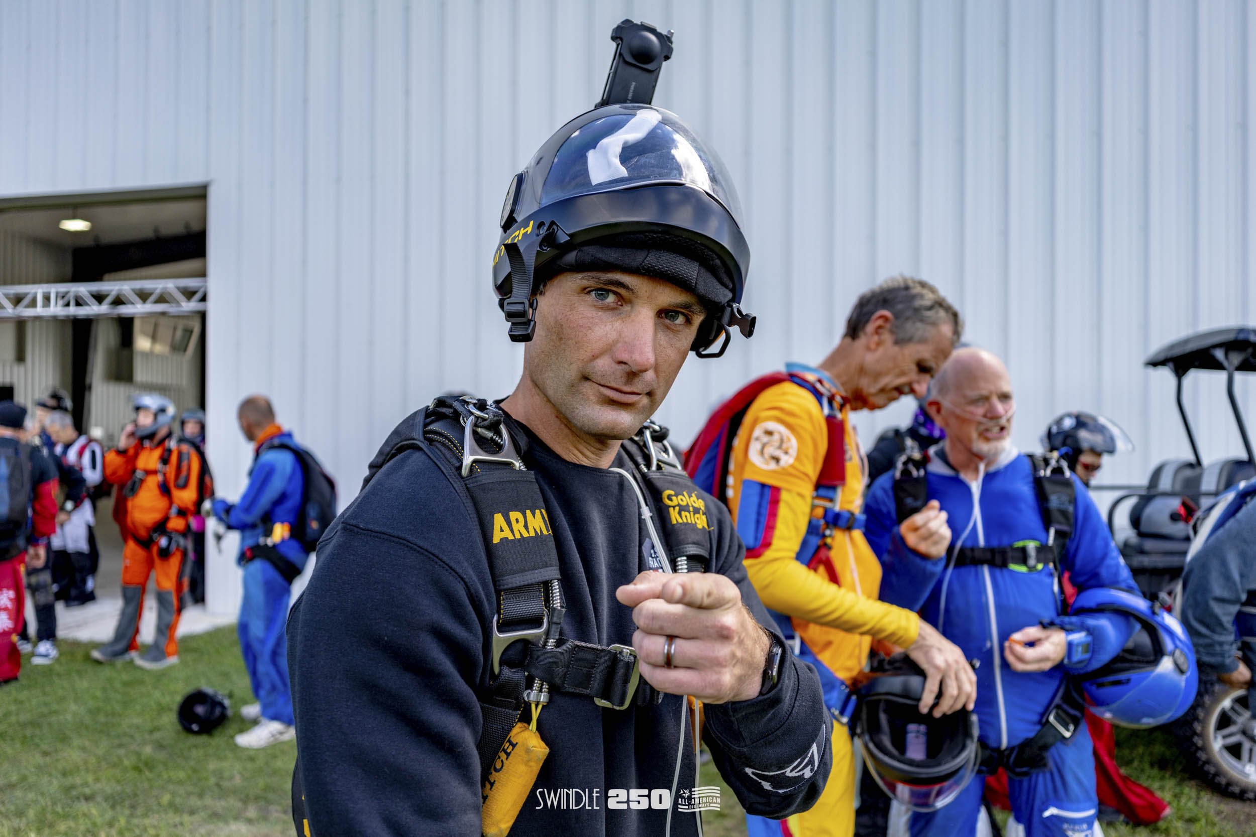 Man in black jumpsuit and helmet posing for camera at skydiving event with others preparing for jump in the background.