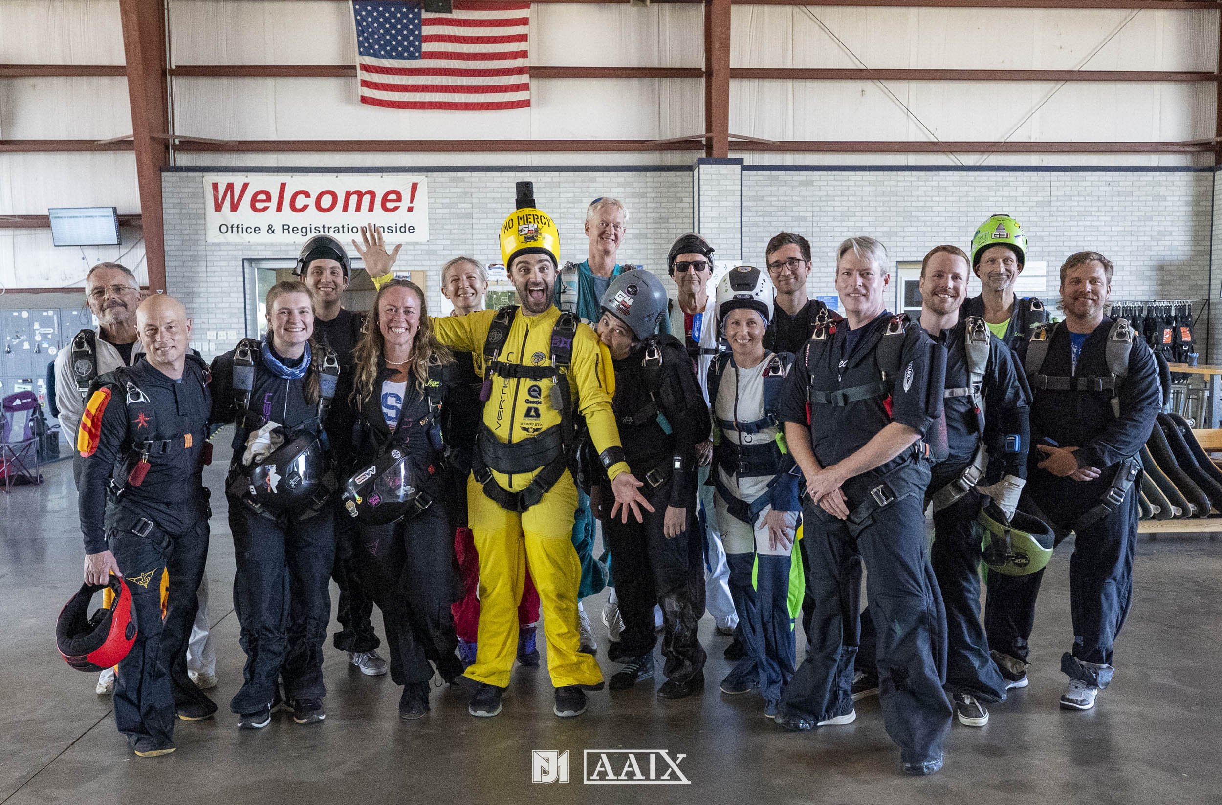 A group of people dressed in skydiving gear, including helmets and harnesses, standing inside a hangar with an American flag and a welcome sign in the background.