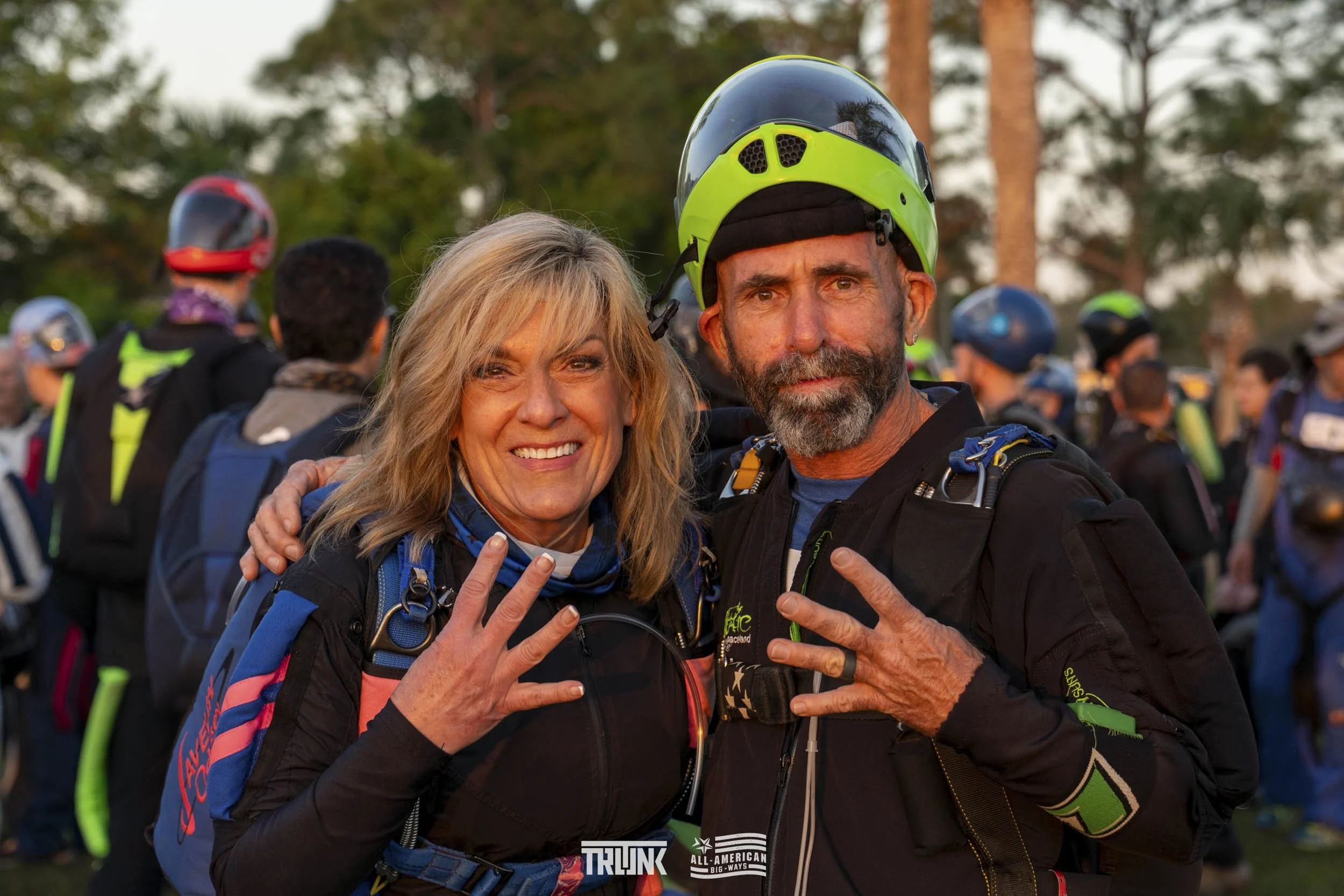 A smiling woman and a serious man wearing motorcycle gear and helmets, making peace signs, at an outdoor event with many people in the background.