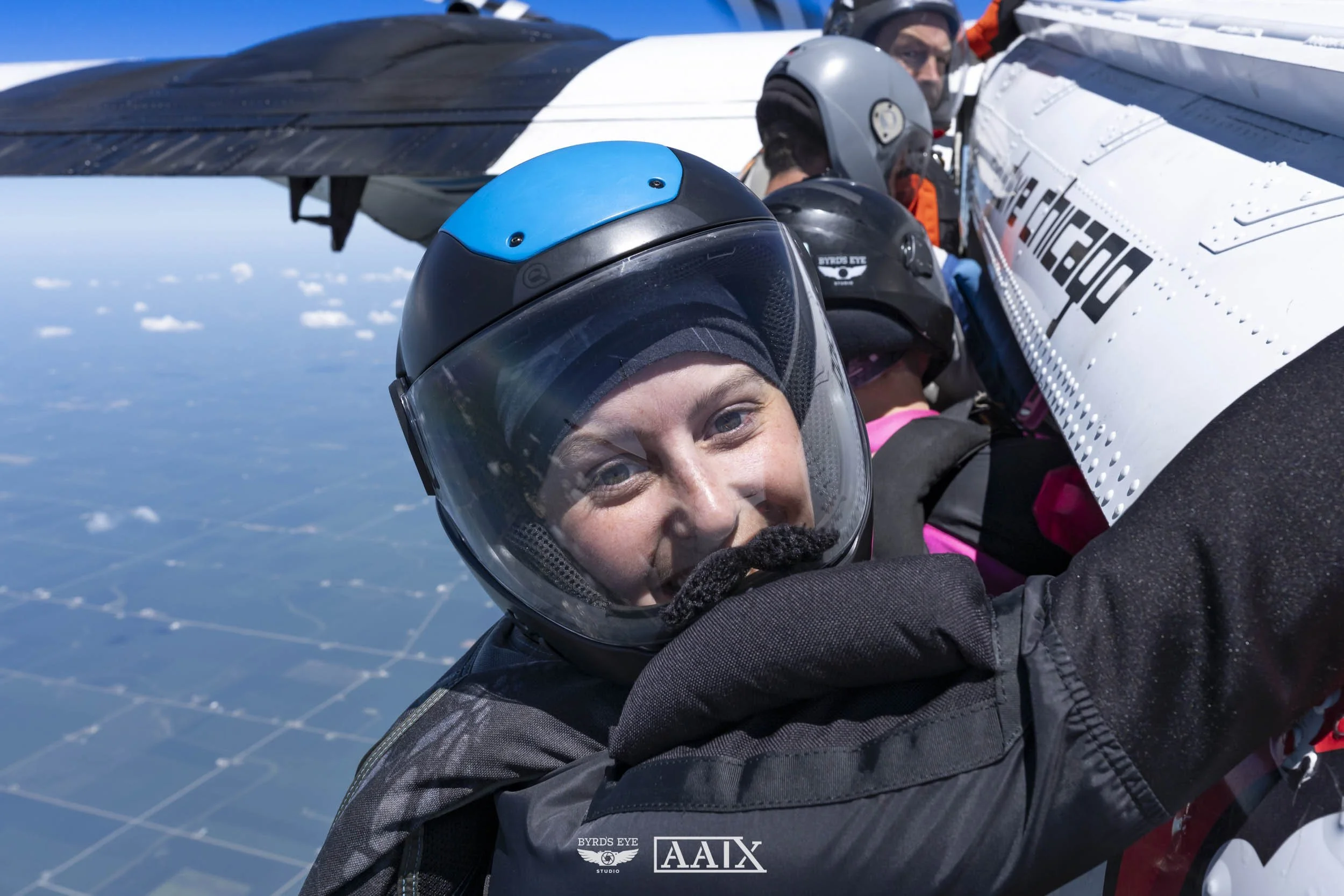 A group of people in helmets and protective gear inside an aircraft, smiling and taking a selfie, with the sky and clouds outside the window.