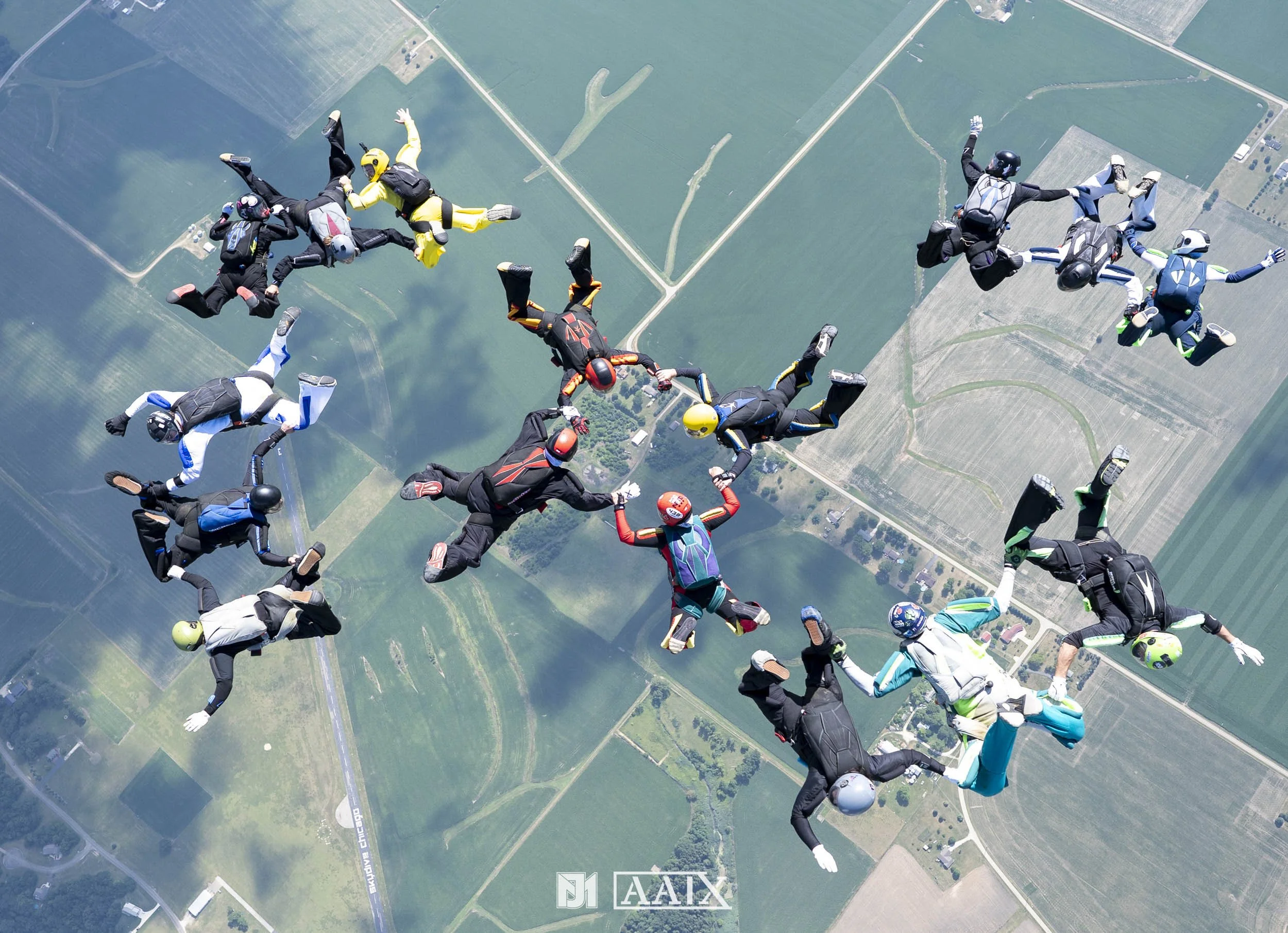 Group of skydivers holding hands in a circle during freefall over green fields and farmland.
