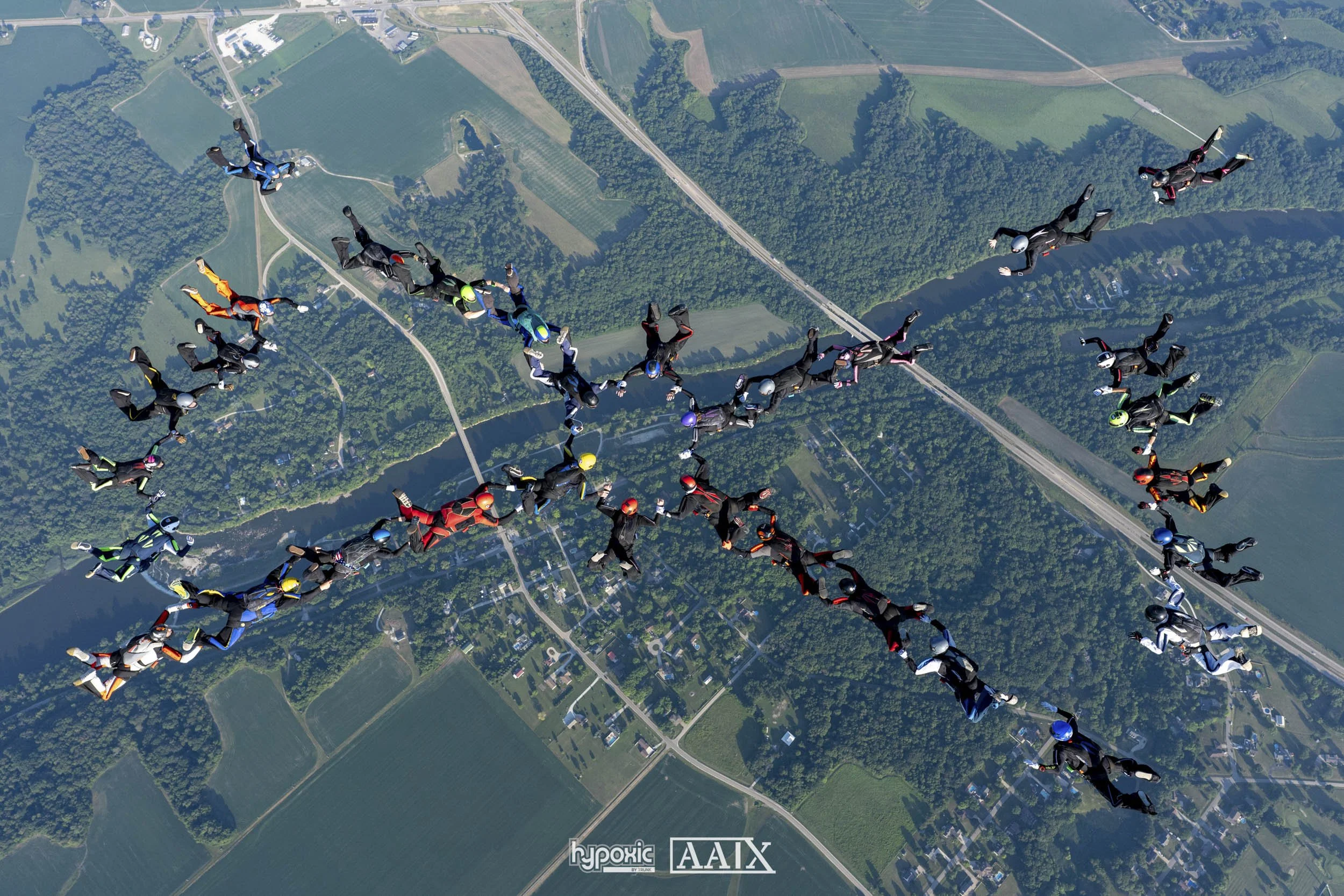 A group of skydivers in free fall over a landscape of fields, forests, and a river, forming the shape of a tree with branches.